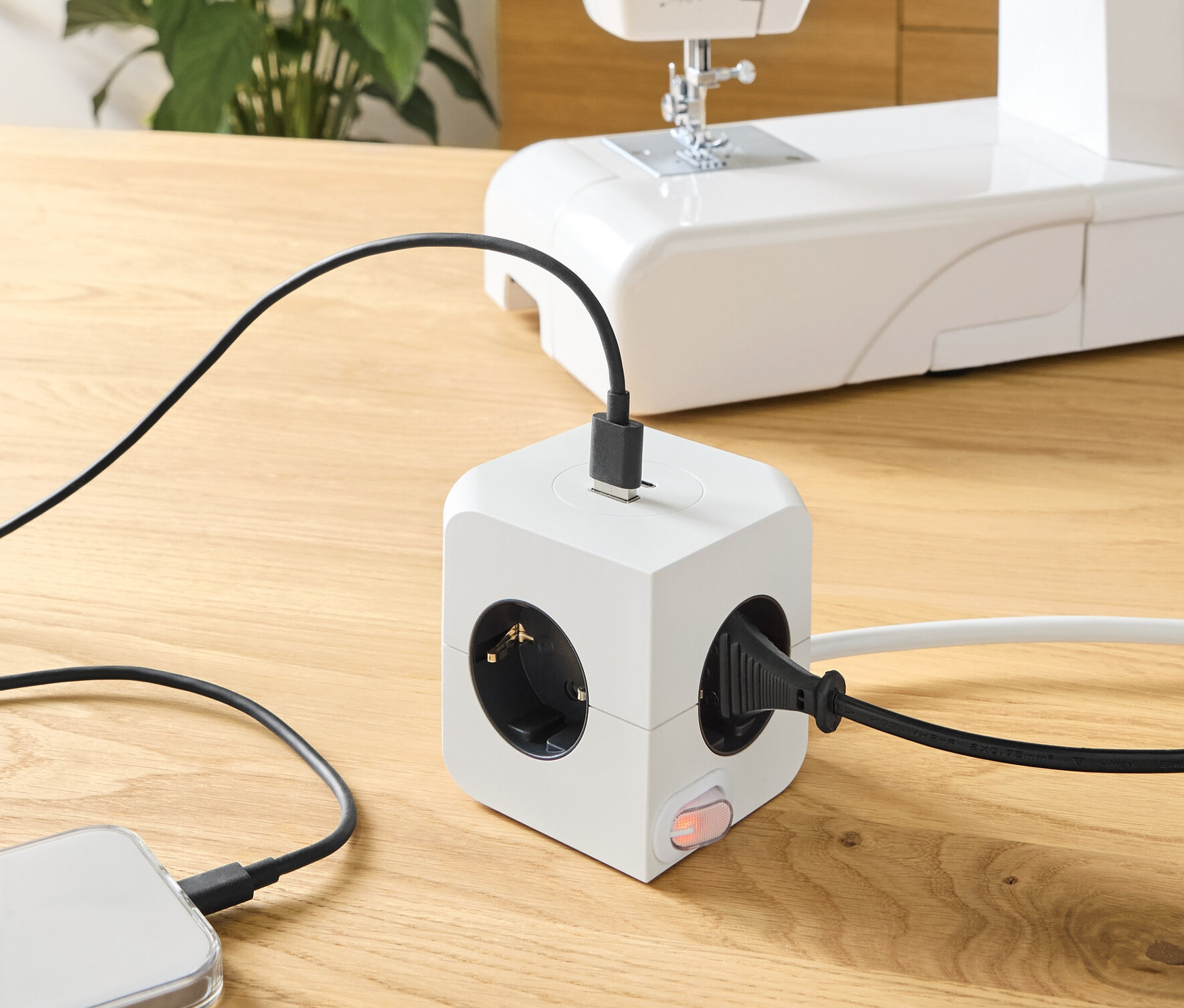 Na dřevěném stole je krychle zásuvky s USB, do které je zapojený telefon a šicí stroj.