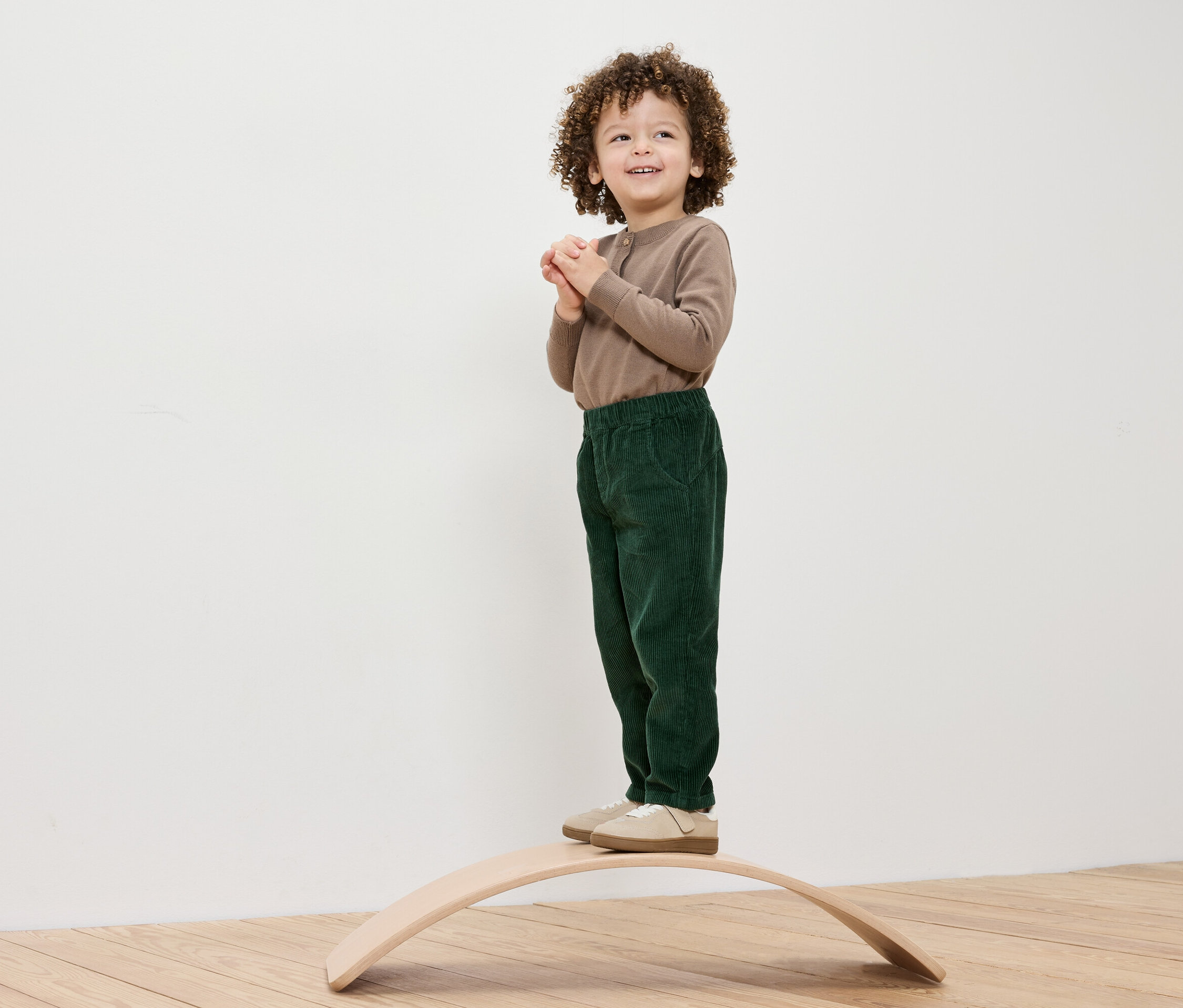 Chlapec s kudrnatými vlasy stojí na dřevěné balanční desce, má na sobě hnědé tričko a zelené kalhoty.