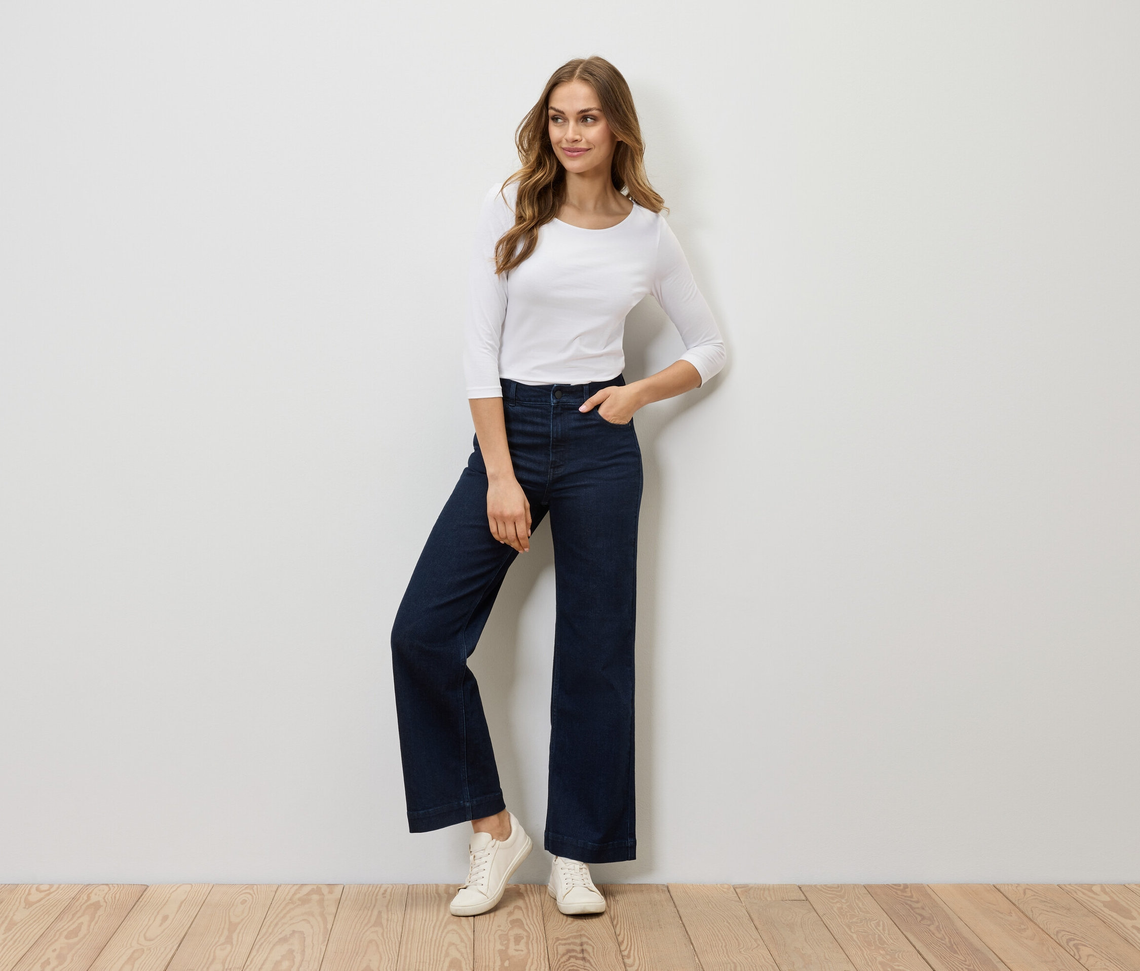 Žena pózuje v bílém tričku s tříčtvrtečním rukávem, širokých tmavě modrých džínách a bílých teniskách.