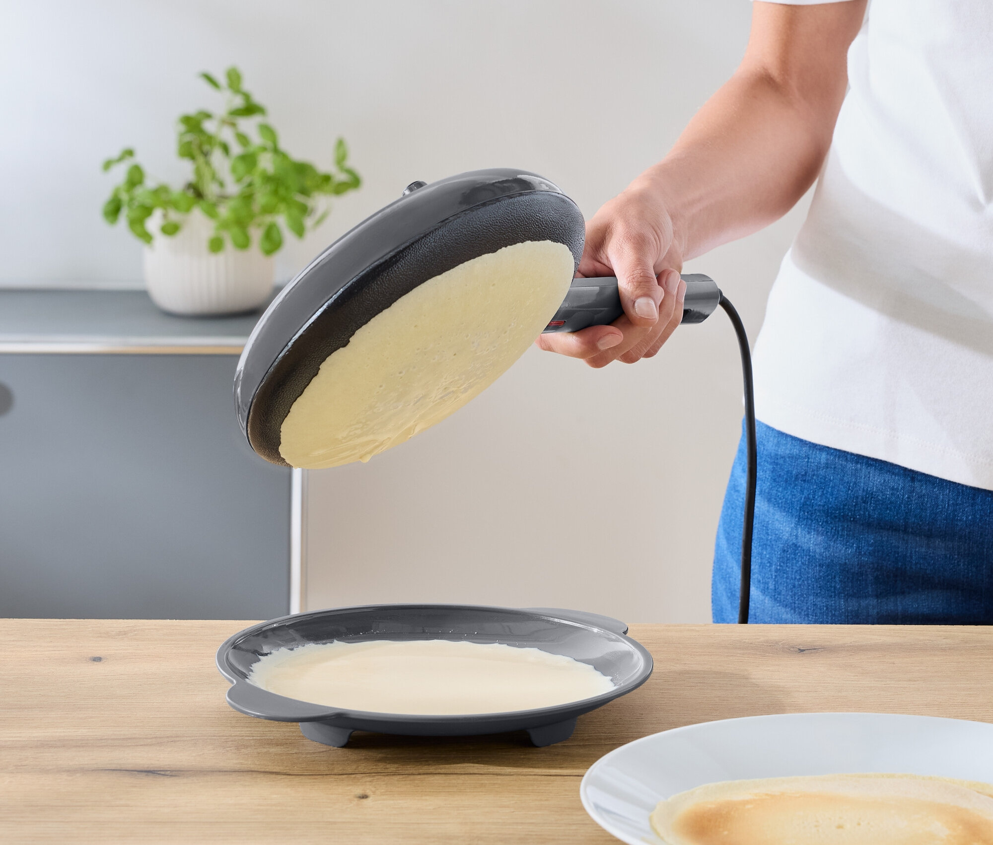 Osoba naklání tmavě šedou elektrickou pánev nad talíř se zbytkem těsta. Hotová palačinka leží na bílém talíři na dřevěné lince.