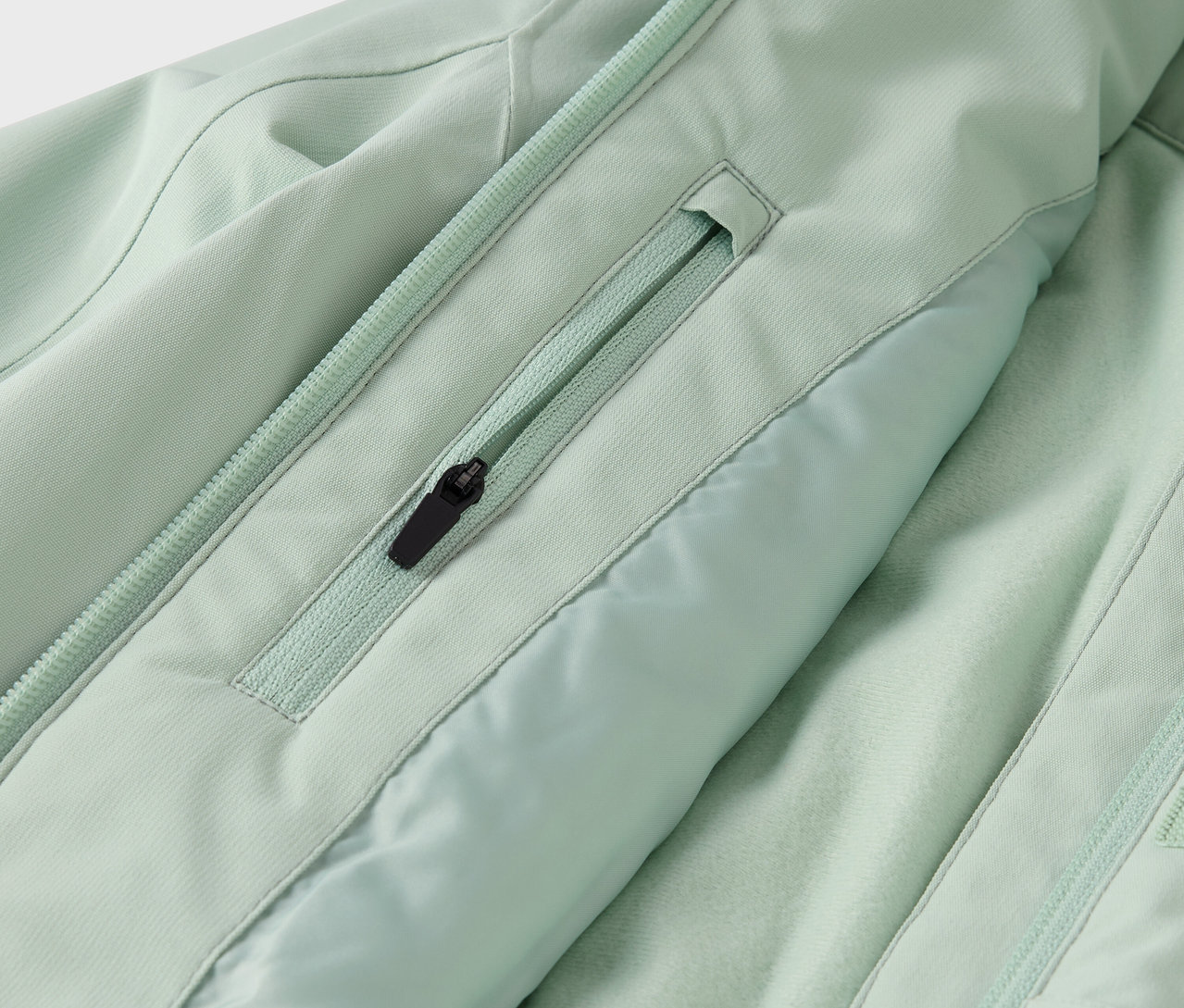 Detail světle zelené bundy s kapsou na zip a zipem uprostřed.