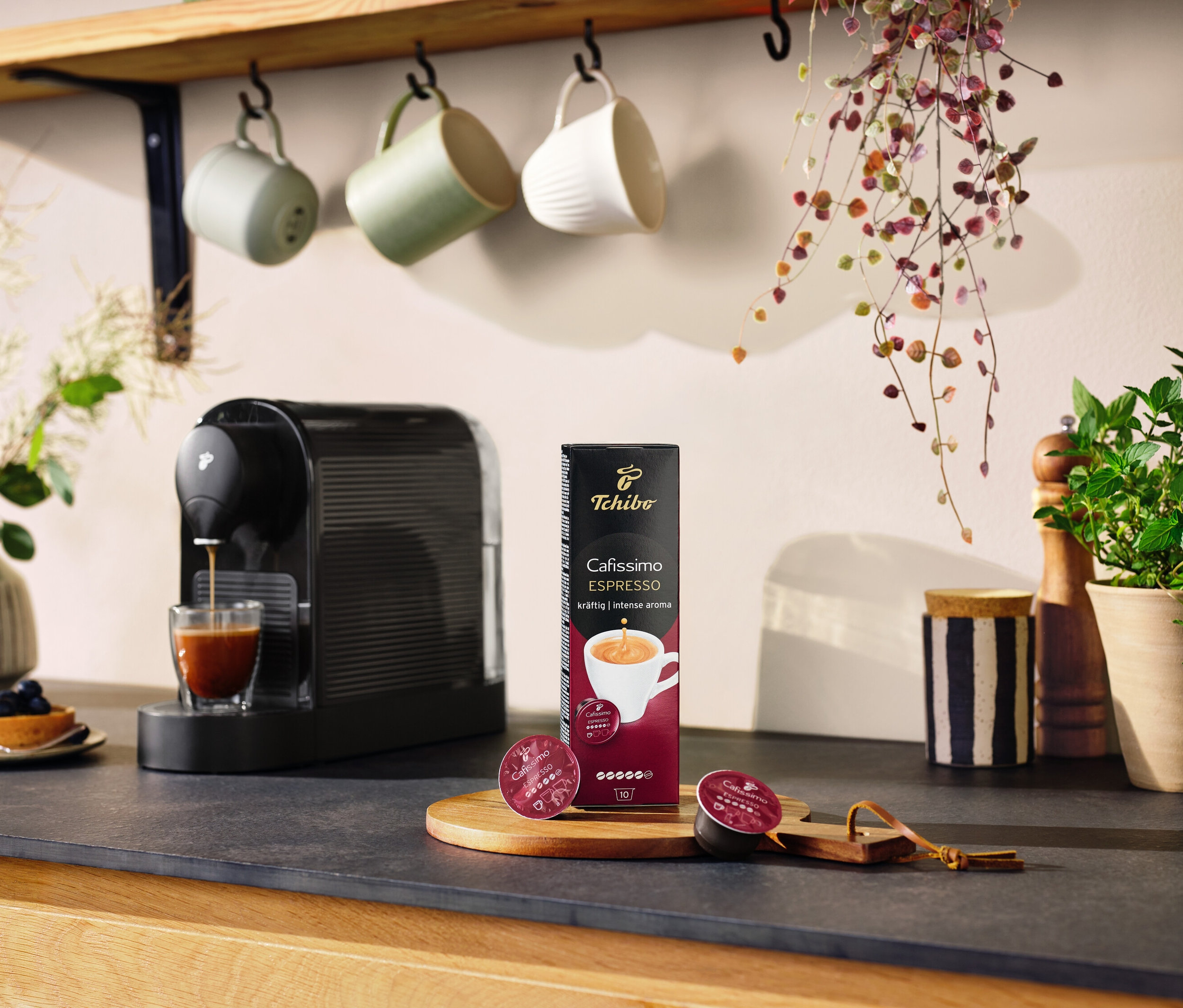 Černý kávovar Cafissimo pure+ připravuje kávu do sklenice. Na stole leží balení kapslí a dvě kapsle.