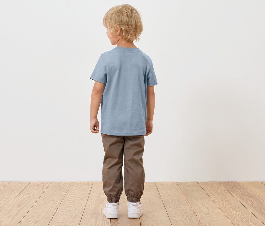 Chlapec s blond vlasy stojí zády, má na sobě modré tričko, hnědé kalhoty a bílé boty.