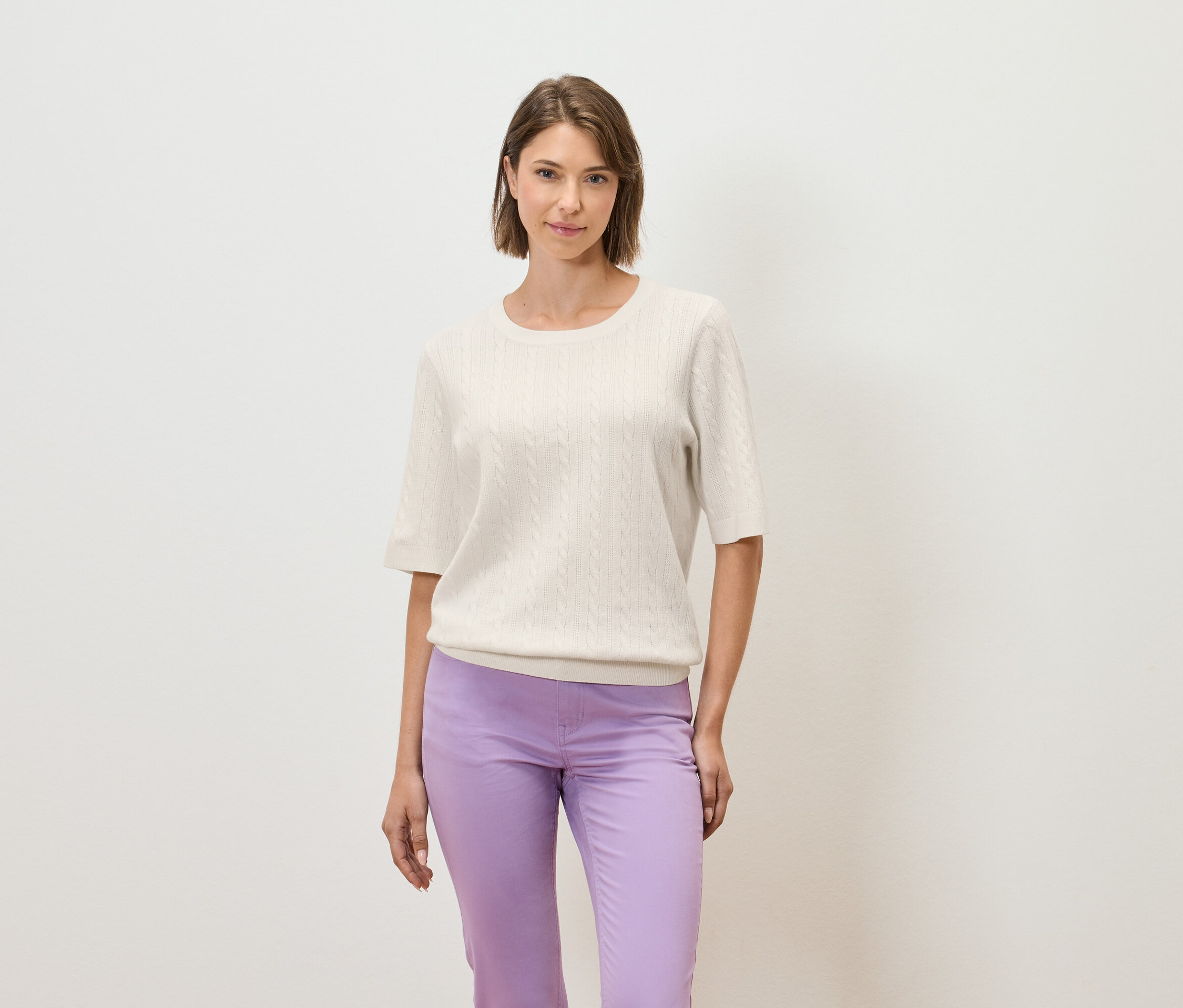 Žena pózuje na světlém pozadí v krémovém svetru s krátkým rukávem a fialových kalhotách.
