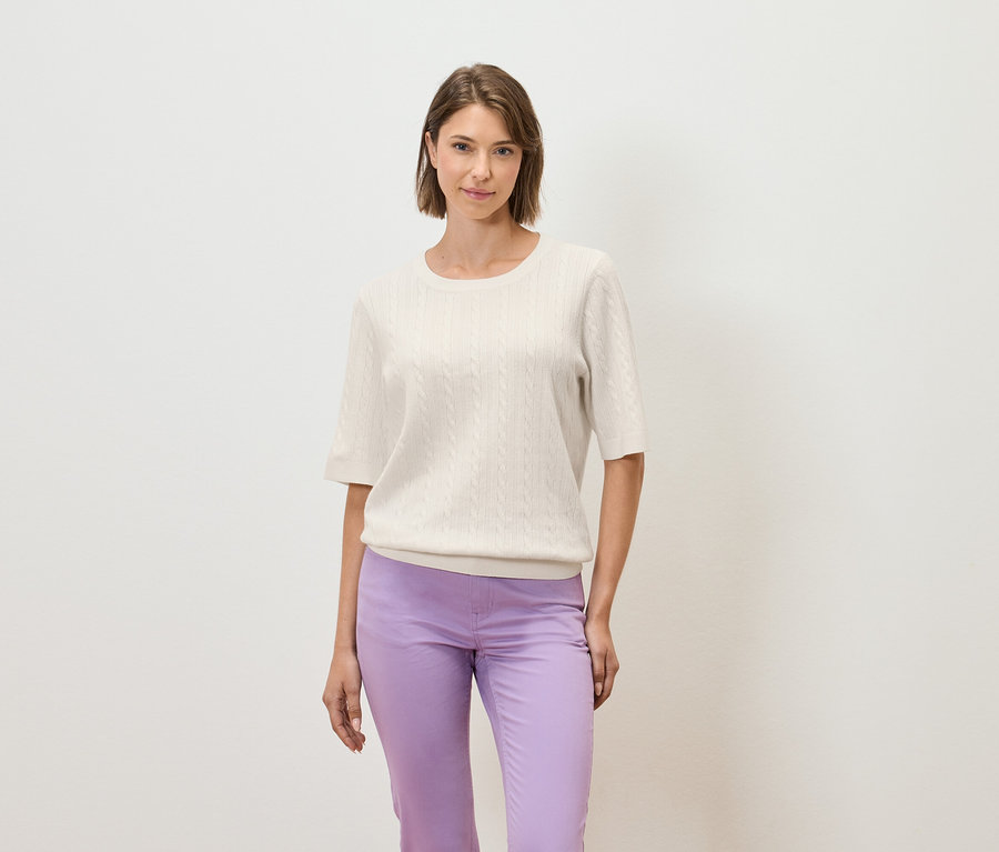 Žena pózuje na světlém pozadí v krémovém svetru s krátkým rukávem a fialových kalhotách.