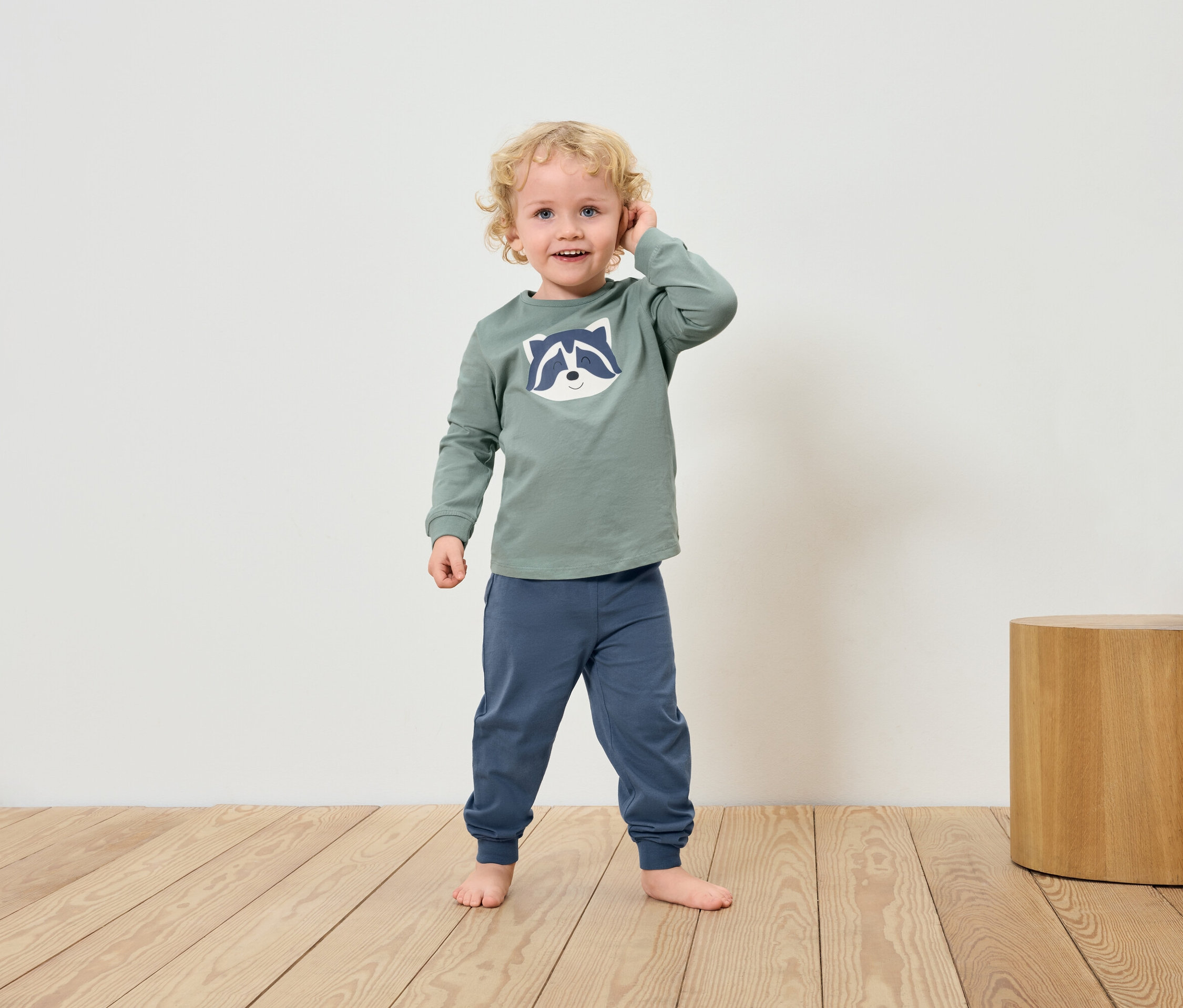 Chlapec s blonďatými vlasy v zeleném tričku s mývalem a modrých kalhotách stojí bosý na dřevěné podlaze.