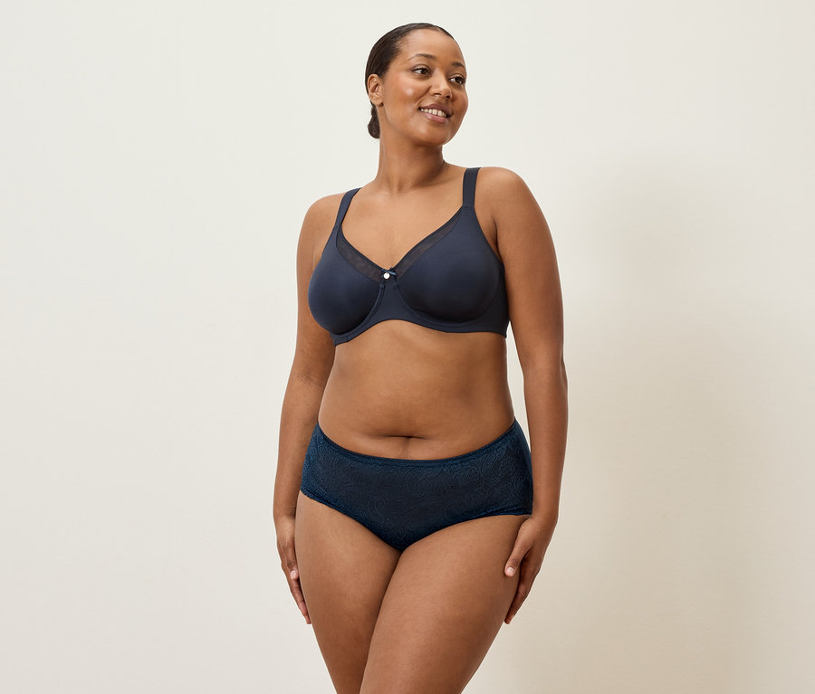 Žena středního věku s tmavými vlasy a tmavě modrým spodním prádlem stojí na krémovém pozadí.