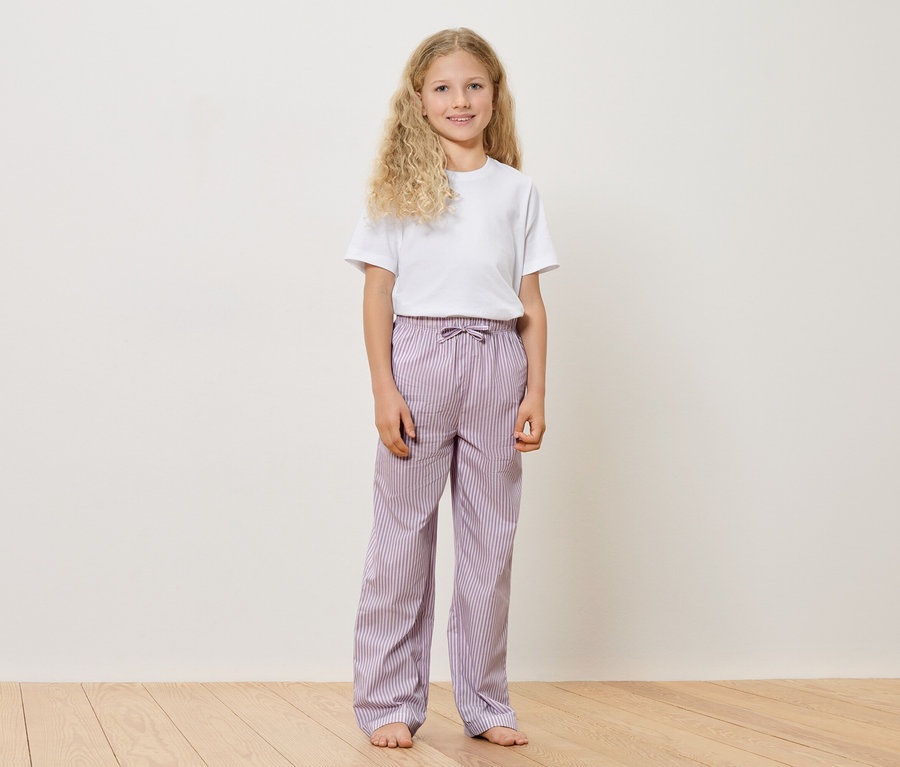 Dívka s blond kudrnatými vlasy v bílém tričku a fialových pruhovaných kalhotách stojí bosá na dřevěné podlaze před bílou zdí.