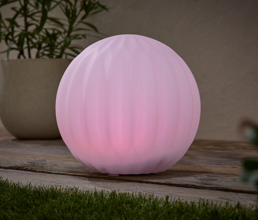 Na dřevěném stole stojí růžová kulová lampa vedle květináče s rostlinou.