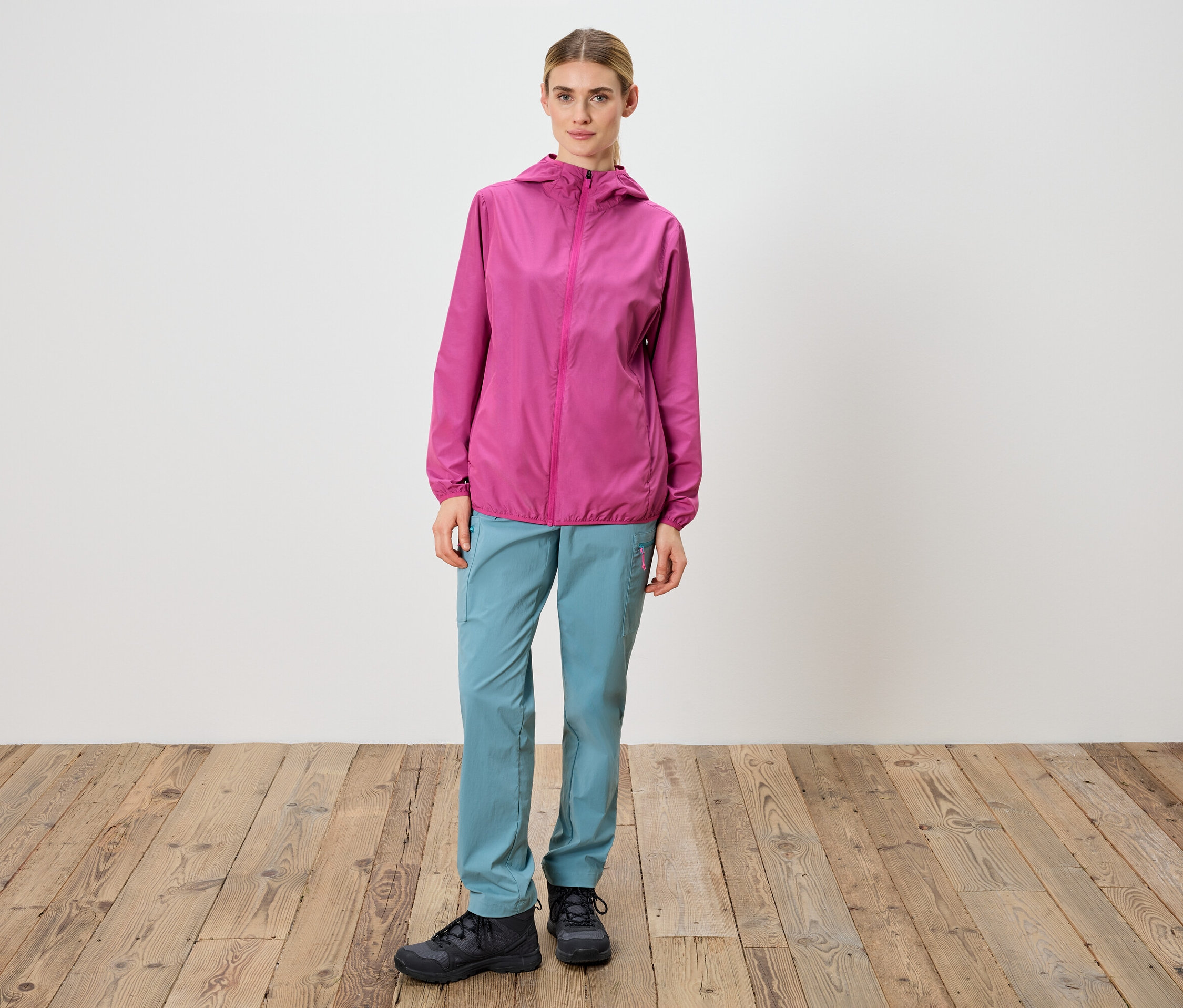 Žena stojí před bílou stěnou a má na sobě růžovou bundu, modré kalhoty a černé boty.