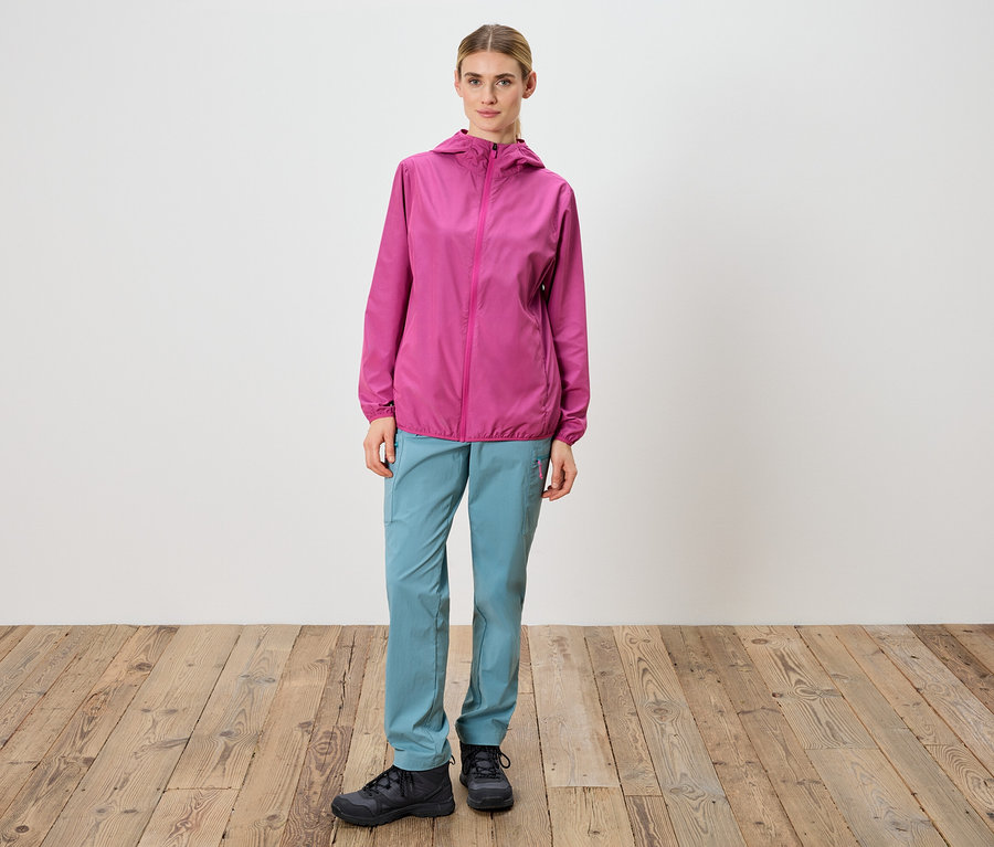 Žena stojí před bílou stěnou a má na sobě růžovou bundu, modré kalhoty a černé boty.