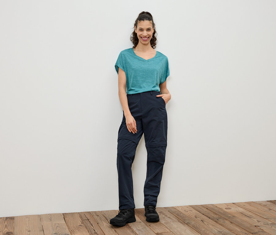 Žena stojí před bílou stěnou a má na sobě modrozelené tričko, tmavě modré kalhoty na zip a černé boty.