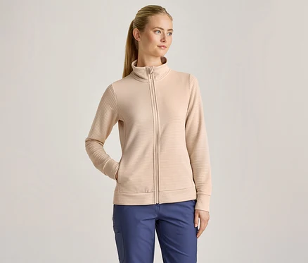 Žena s blond culíkem má na sobě béžovou žebrovanou bundu na zip a modré kalhoty, s jednou rukou v kapse bundy.