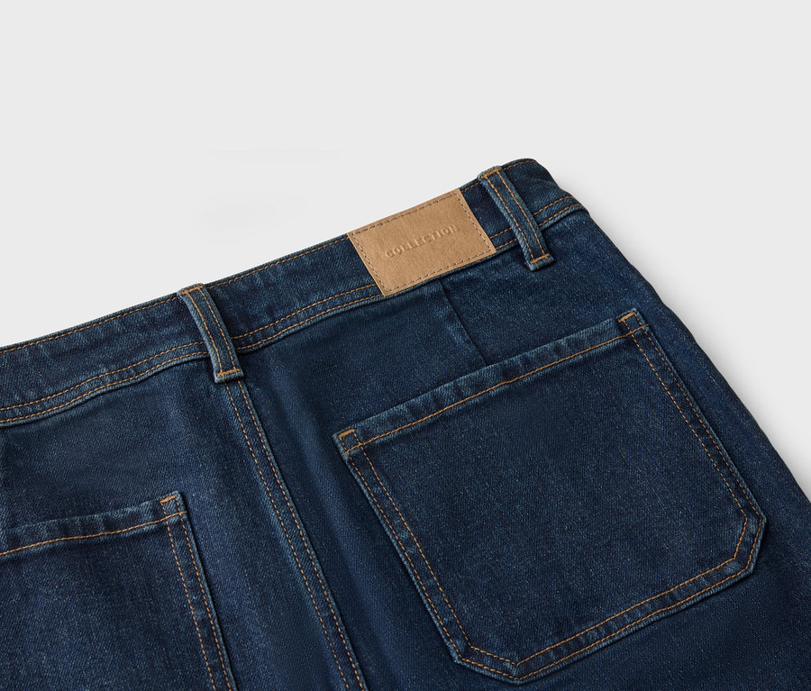 Detailní záběr na zadní část tmavě modrých džínů s našitými kapsami a hnědou koženou nášivkou s nápisem „Collection“.