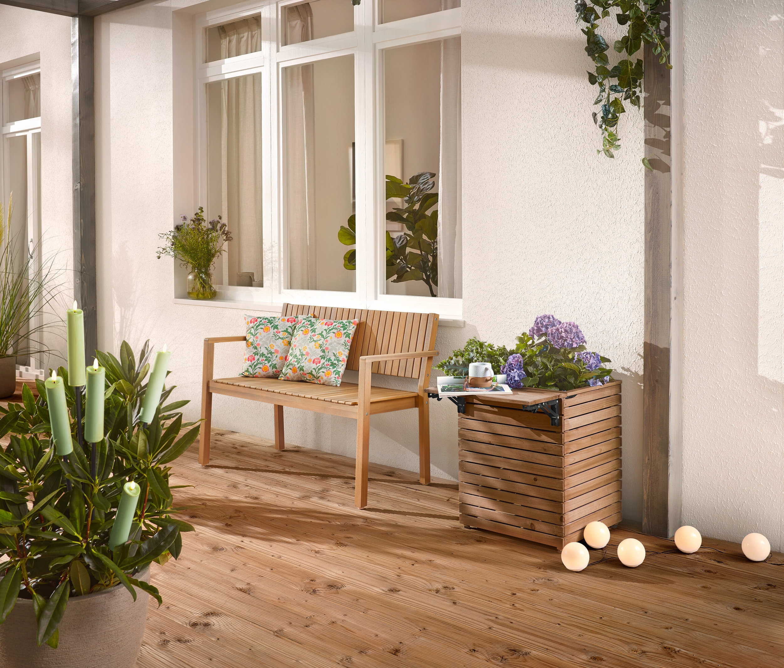 Na dřevěné terase stojí dřevěná lavice s polštáři, truhlík s květinami a hrnková rostlina se svíčkami.