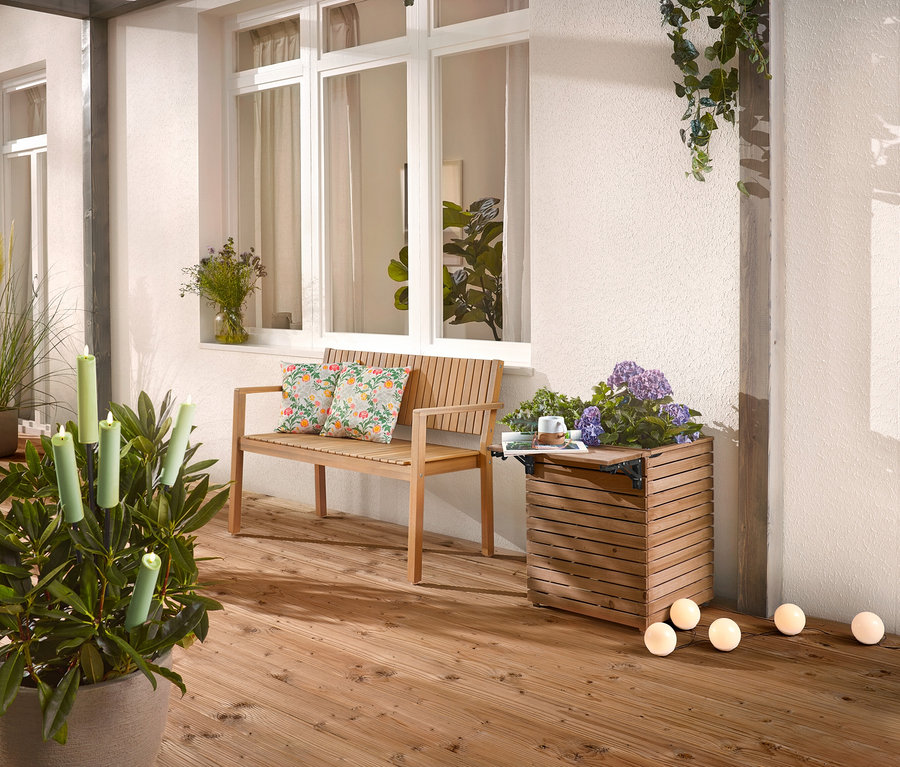 Na dřevěné terase stojí dřevěná lavice s polštáři, truhlík s květinami a hrnková rostlina se svíčkami.