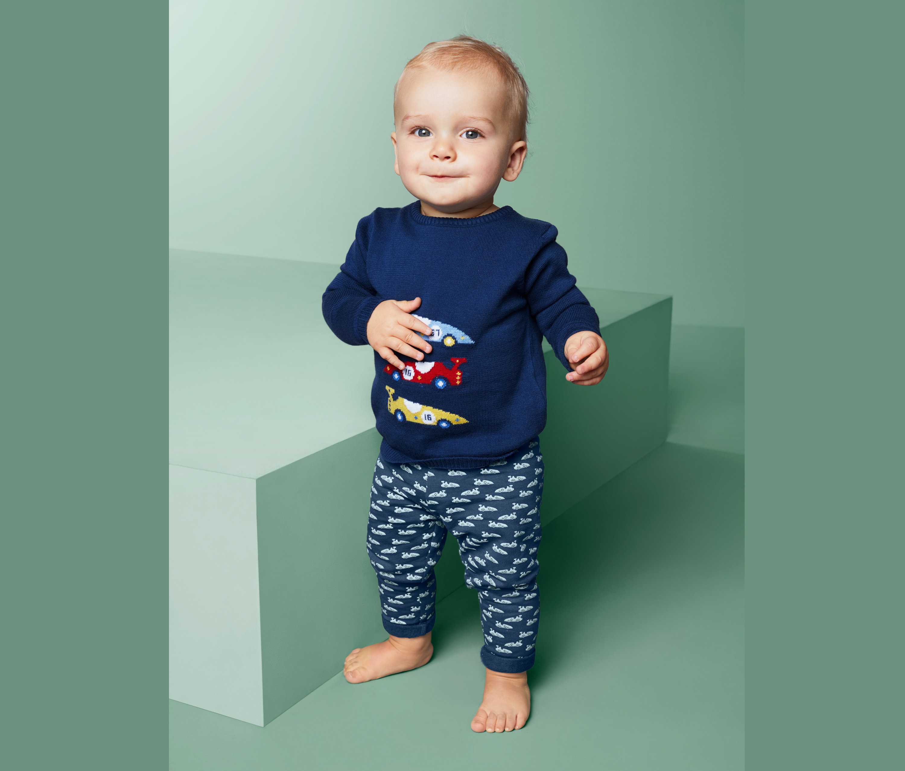 Dítě stojí a má na sobě modrý svetr s auty a teplákové kalhoty.