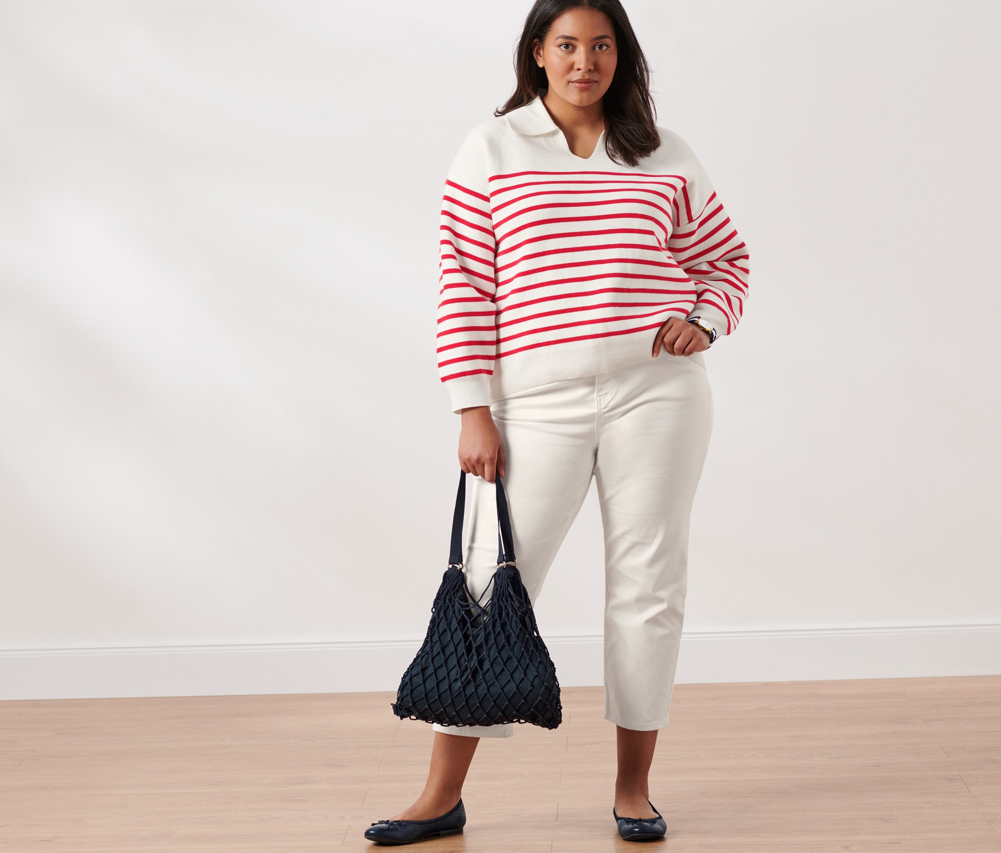 Žena pózuje ve svetru s límcem bílo-červené barvy, džínách slim fit, kožených balerínách, se síťovkou a náramkovými hodinkami z ušlechtilé oceli.