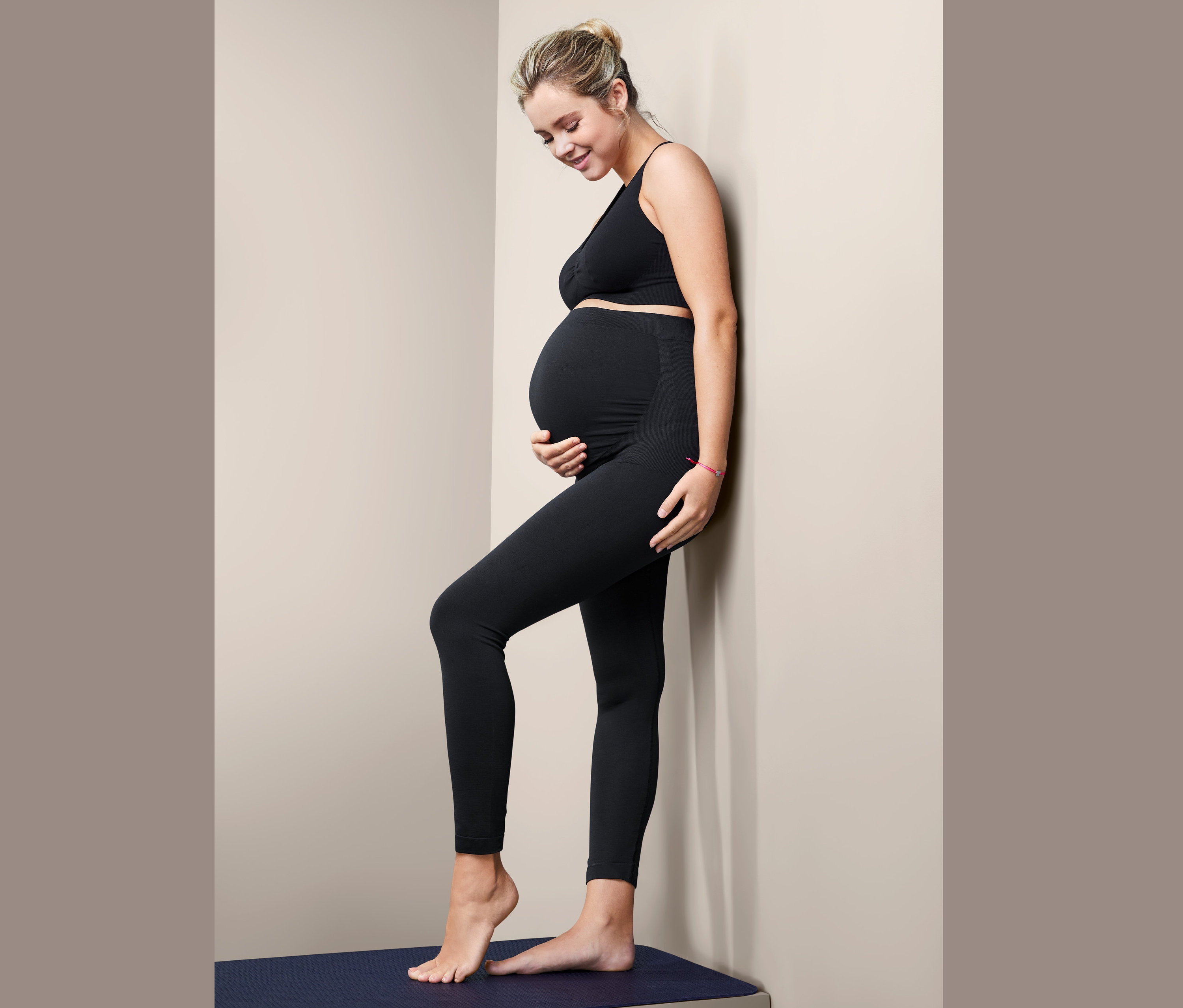 Těhotná žena v černých bezešvých legínách stojí bokem u zdi.