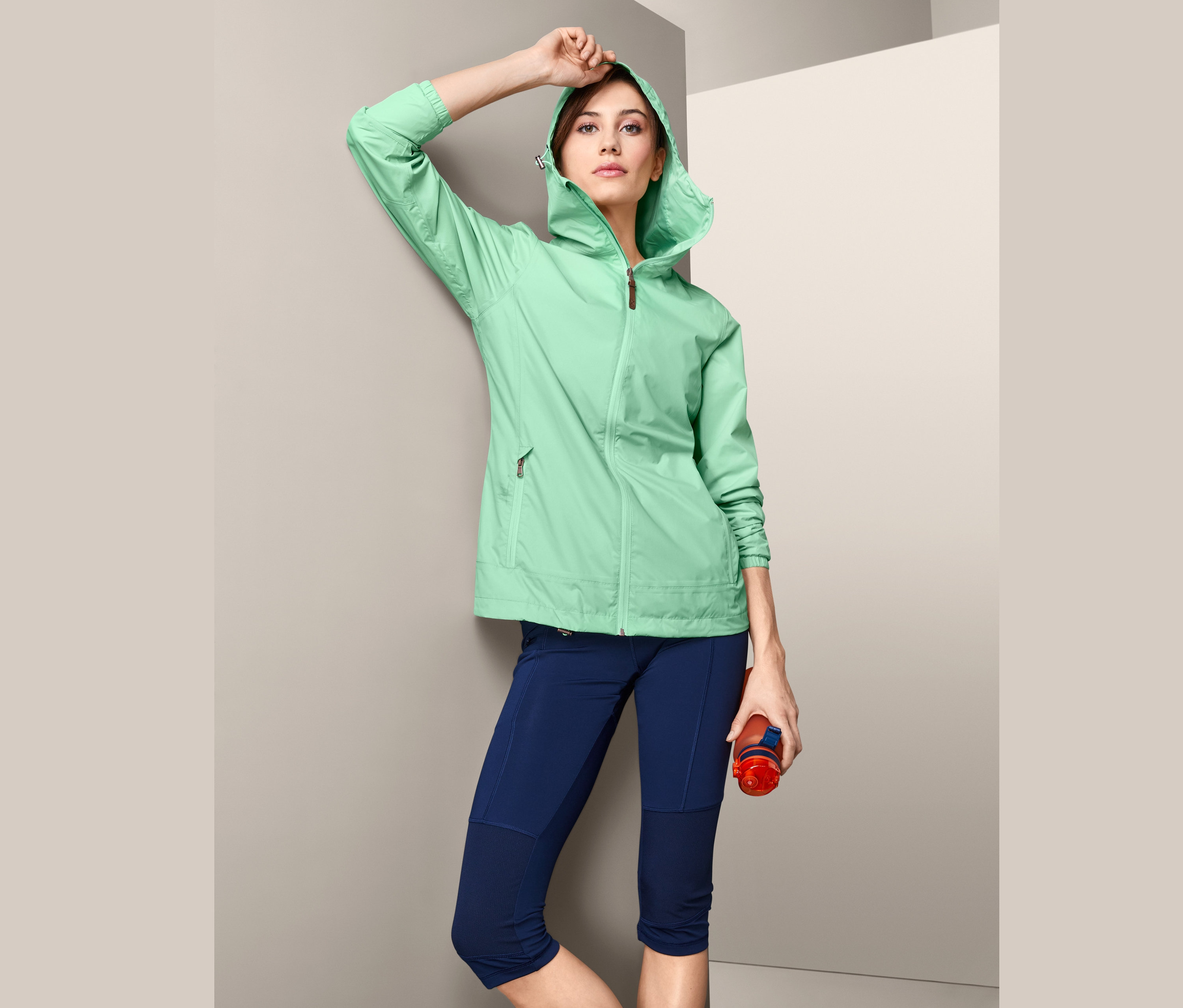 Žena v lehké bundě do deště Pack Me, s kapucí, pózuje s lahví na vodu.