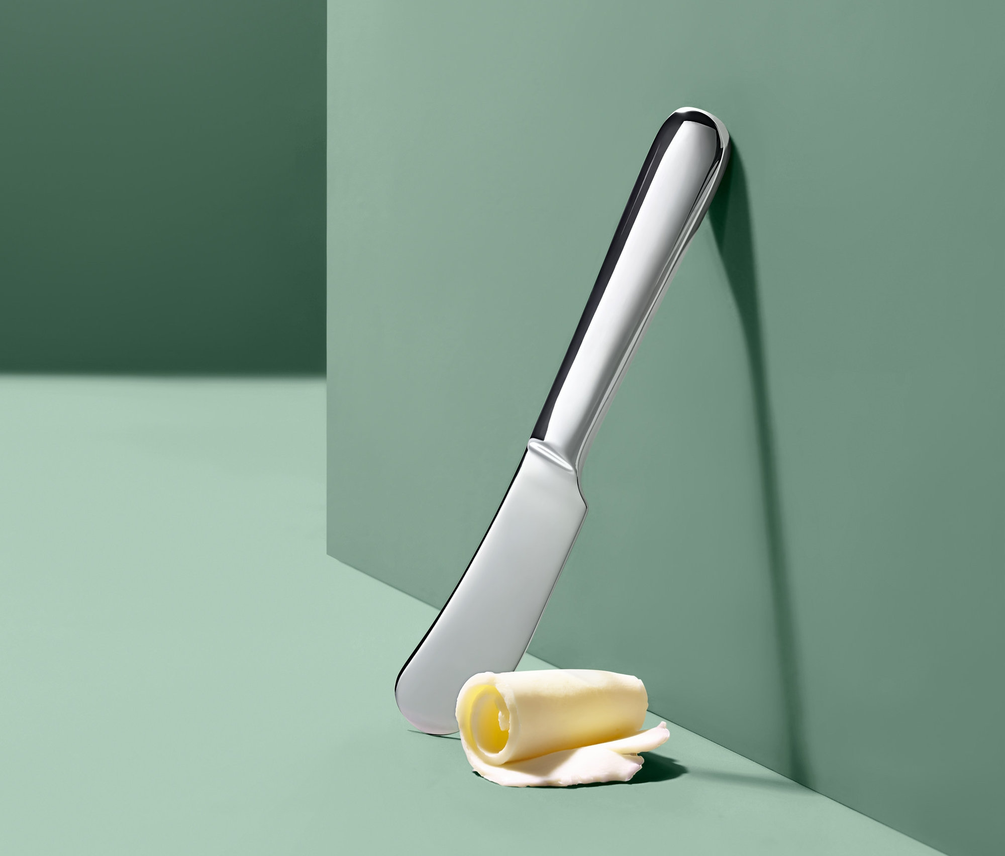 Stříbrný nůž na máslo a rolka másla opřené o zelenou stěnu.