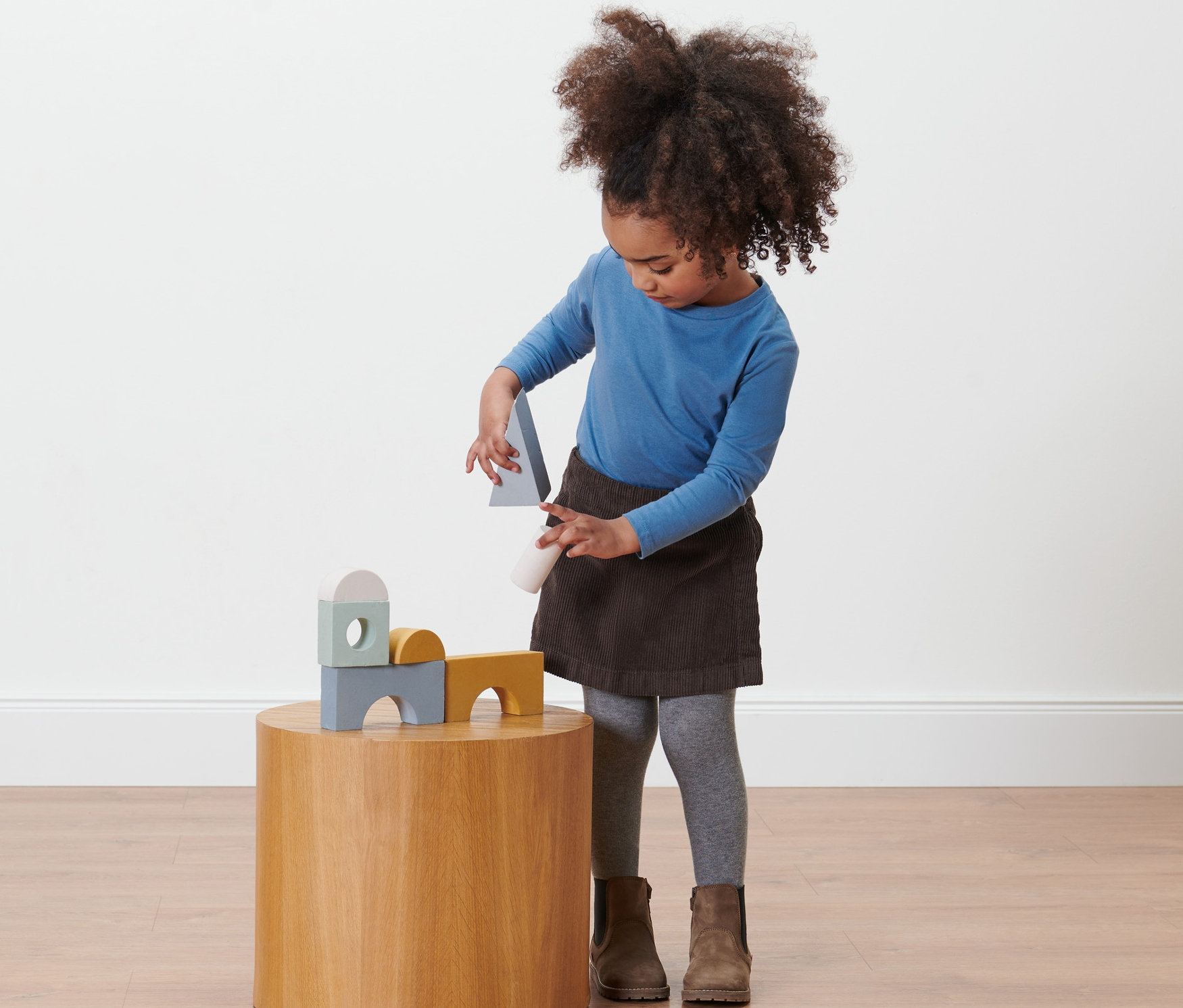 Dívka si hraje s dřevěnými hračkami na kulatém stolku, má na sobě dětskou kalhotovou sukni.