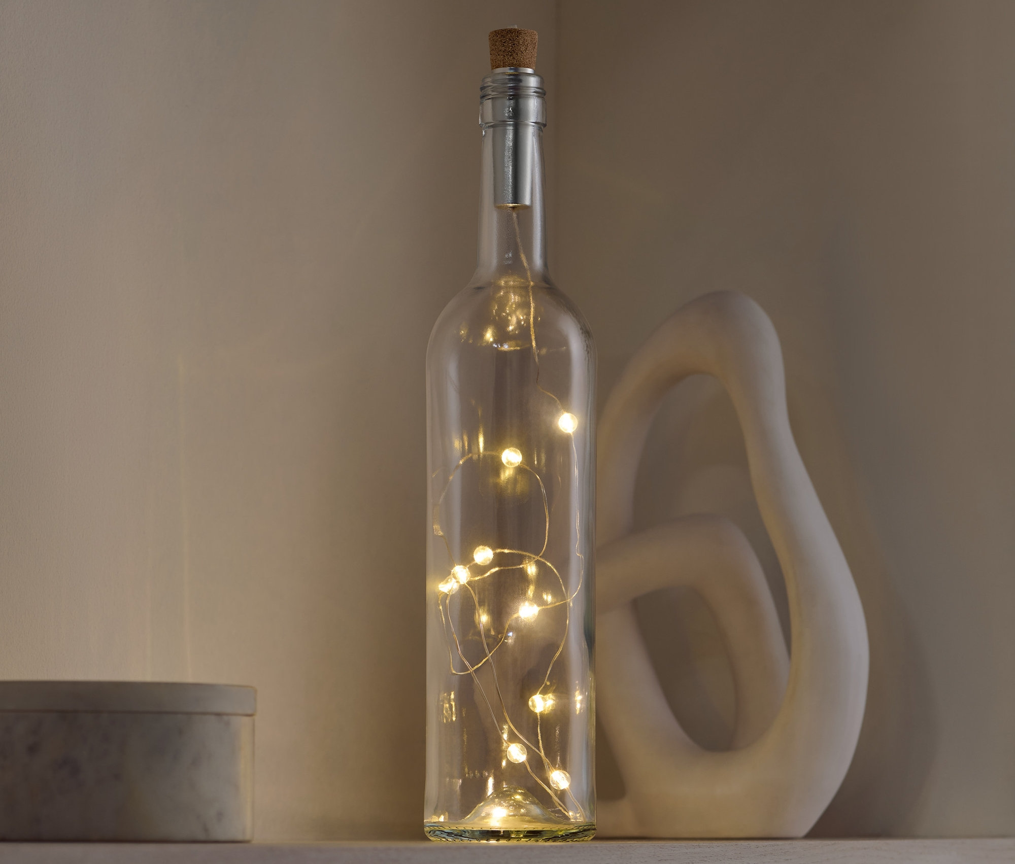 Dekorativní skleněná láhev s korkovou zátkou, uvnitř které jsou světla, stojí na polici vedle ozdobné sošky a šedé krabičky.