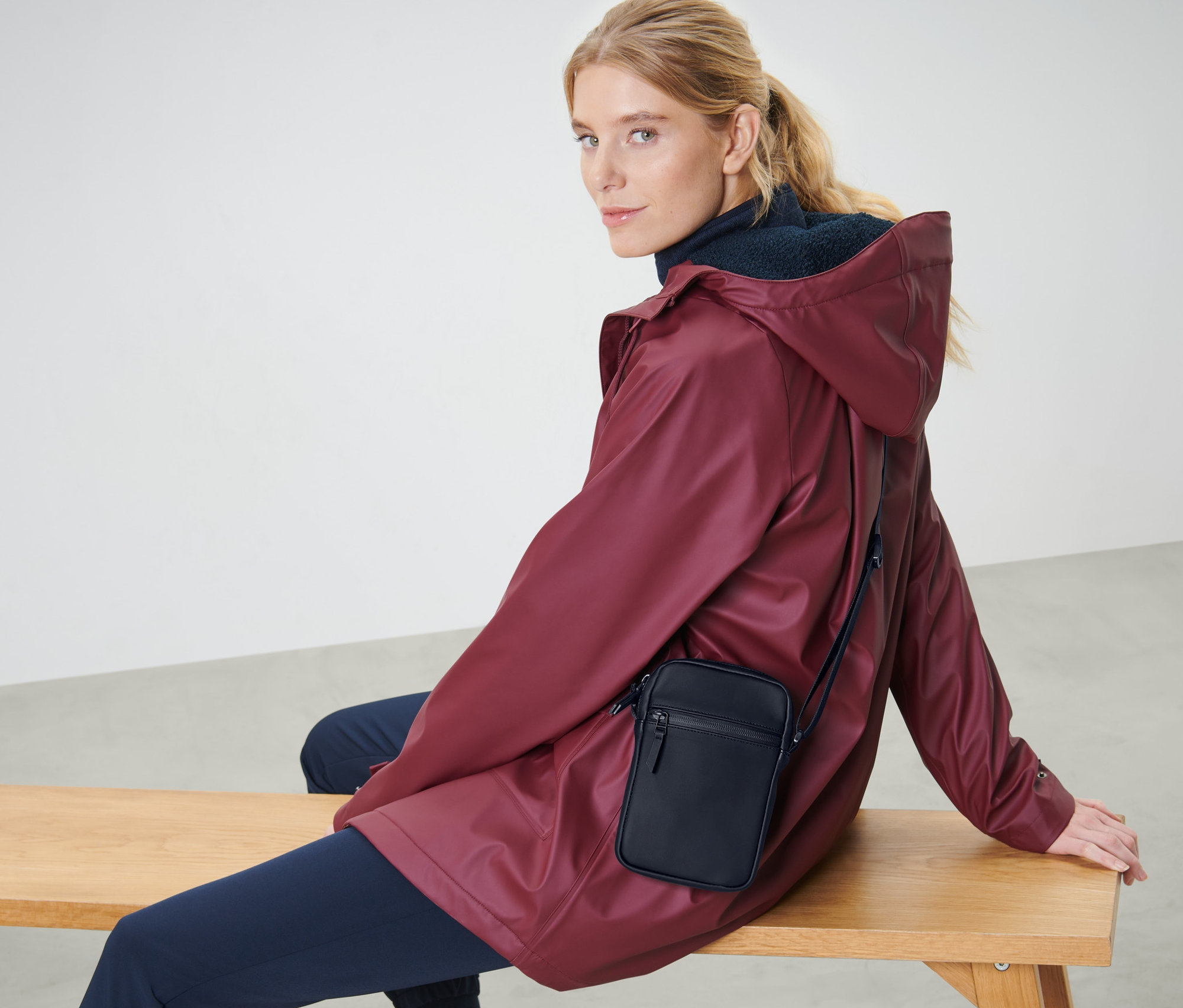 Blondýna sedí na lavici v bordó bundě a s černou taškou přes rameno.