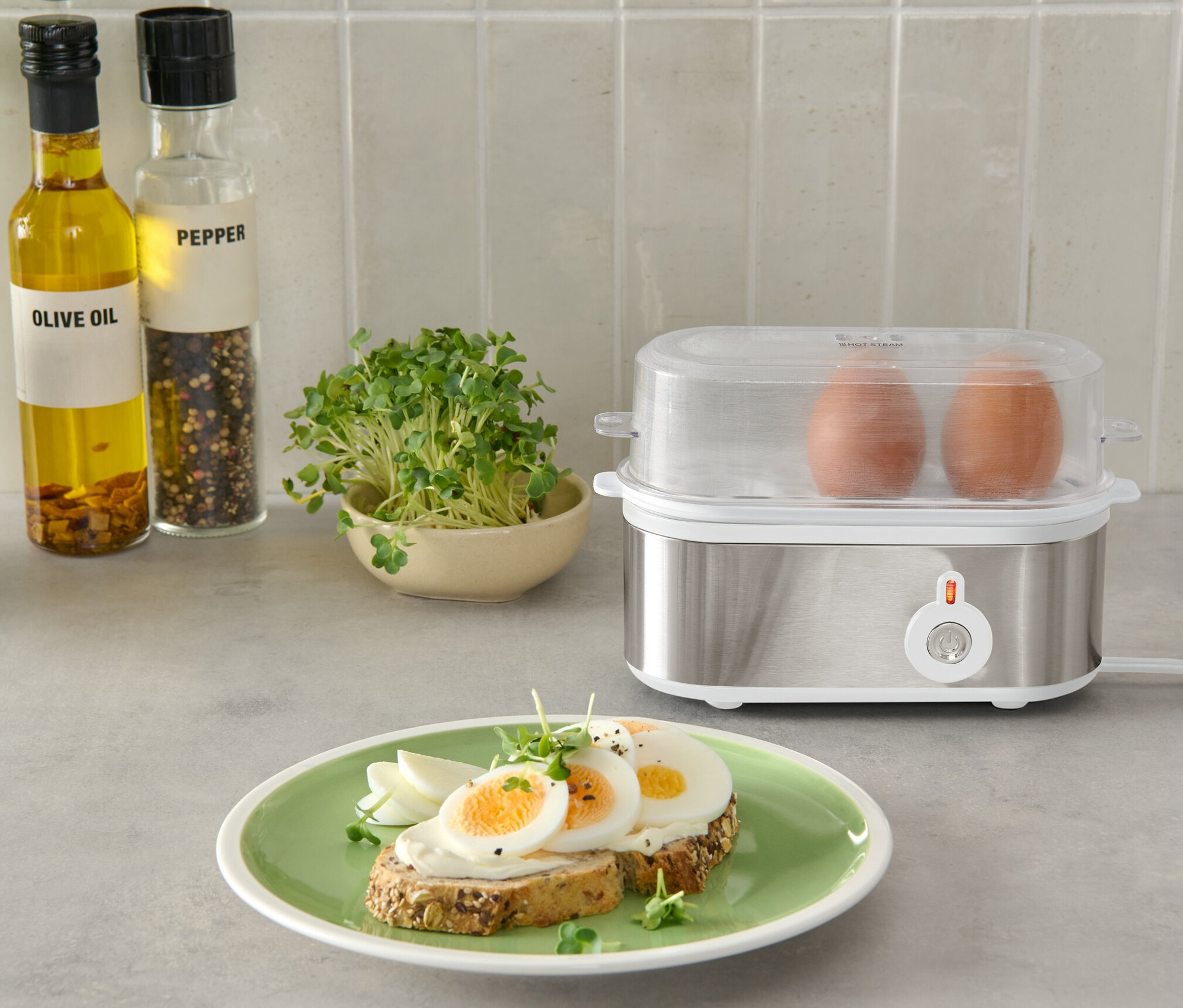 Dva hnědá vejce ve stříbrném vařiči vajec s průhledným víkem, vedle talíře s chlebem a vejci.