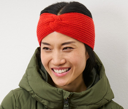 Žena se zkříženýma rukama usmívá se a má na sobě oranžovou pletenou čelenku a zelenou péřovou bundu.