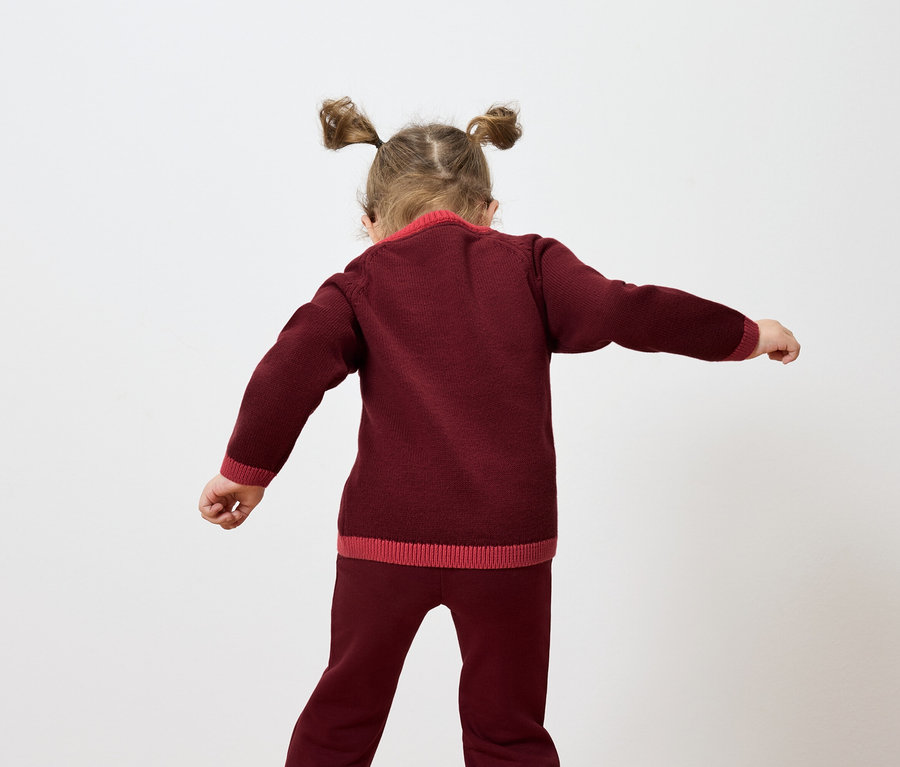 Dívka s culíky stojí zády a mává rukama, oblečená do bordó pleteného svetru a kalhot.