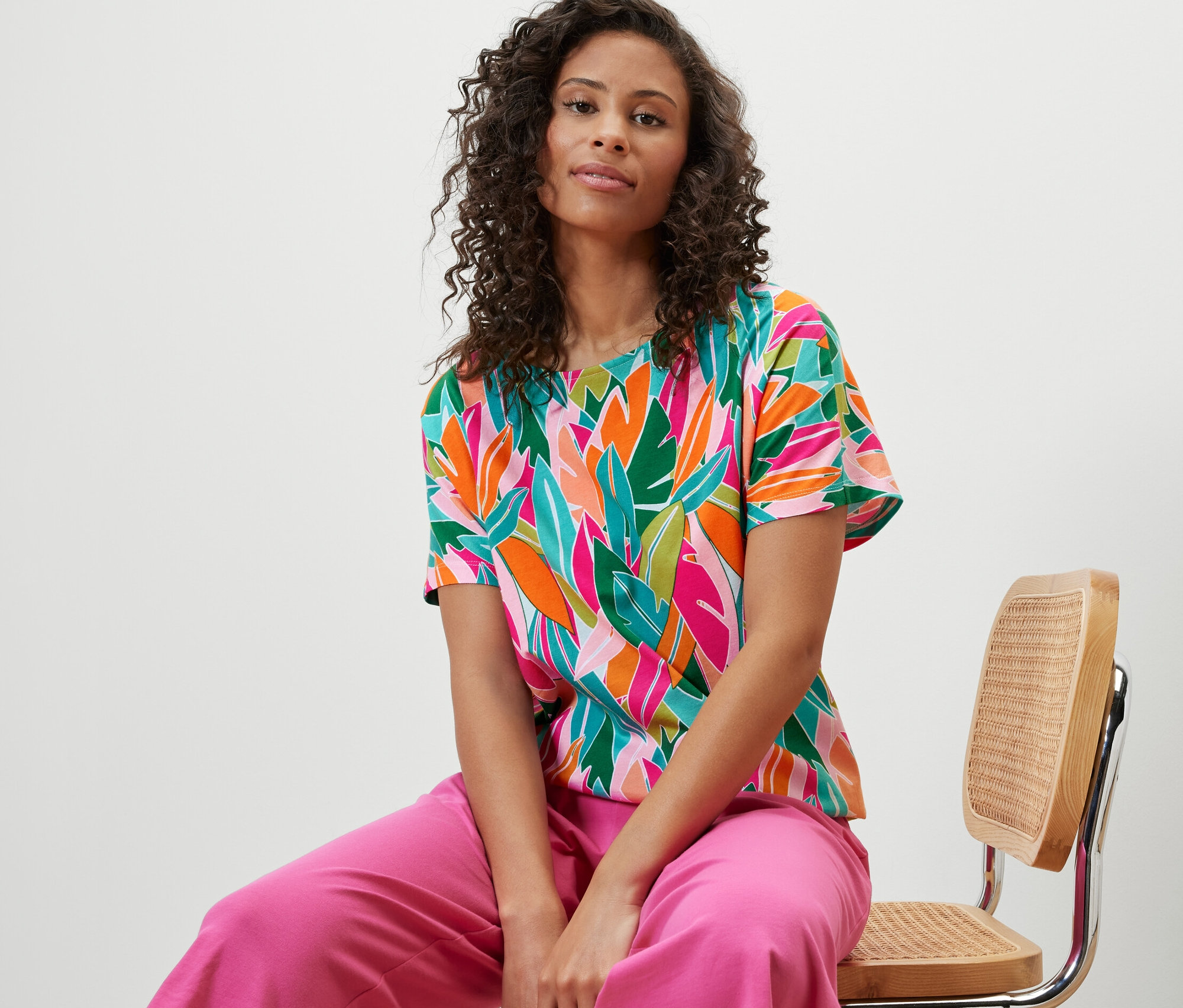 Žena sedí na židli a má na sobě halenkové tričko se spadlými rameny a celoplošným potiskem.