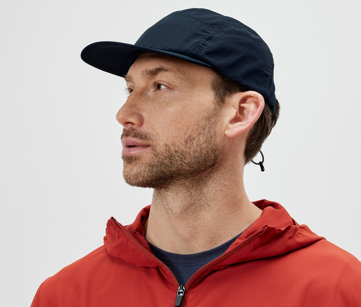 Muž v oranžové bundě má na sobě sbalitelnou kšiltovku a dívá se doleva.