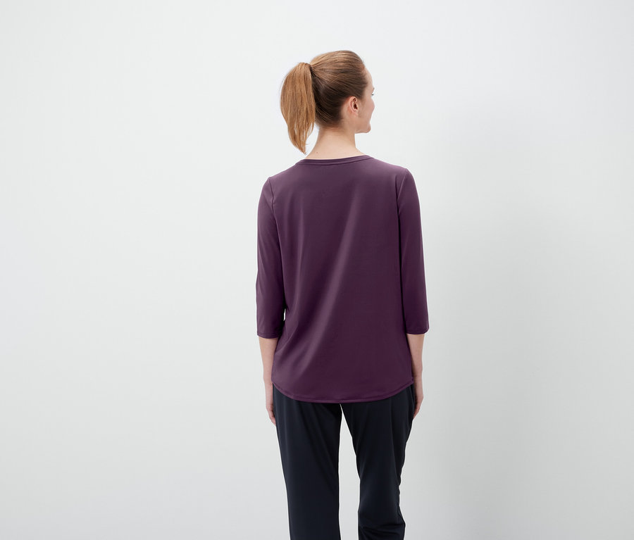 Zezadu pohled na ženu s hnědými vlasy staženými do culíku, oblečenou do fialového trička s tříčtvrtečními rukávy a černých kalhot.