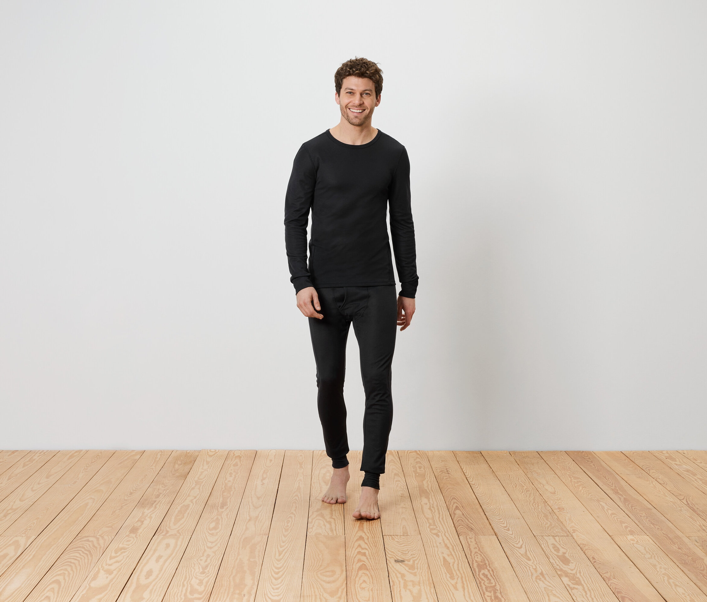 Muž v černém termoprádle stojí bosý na dřevěné podlaze před bílou zdí.