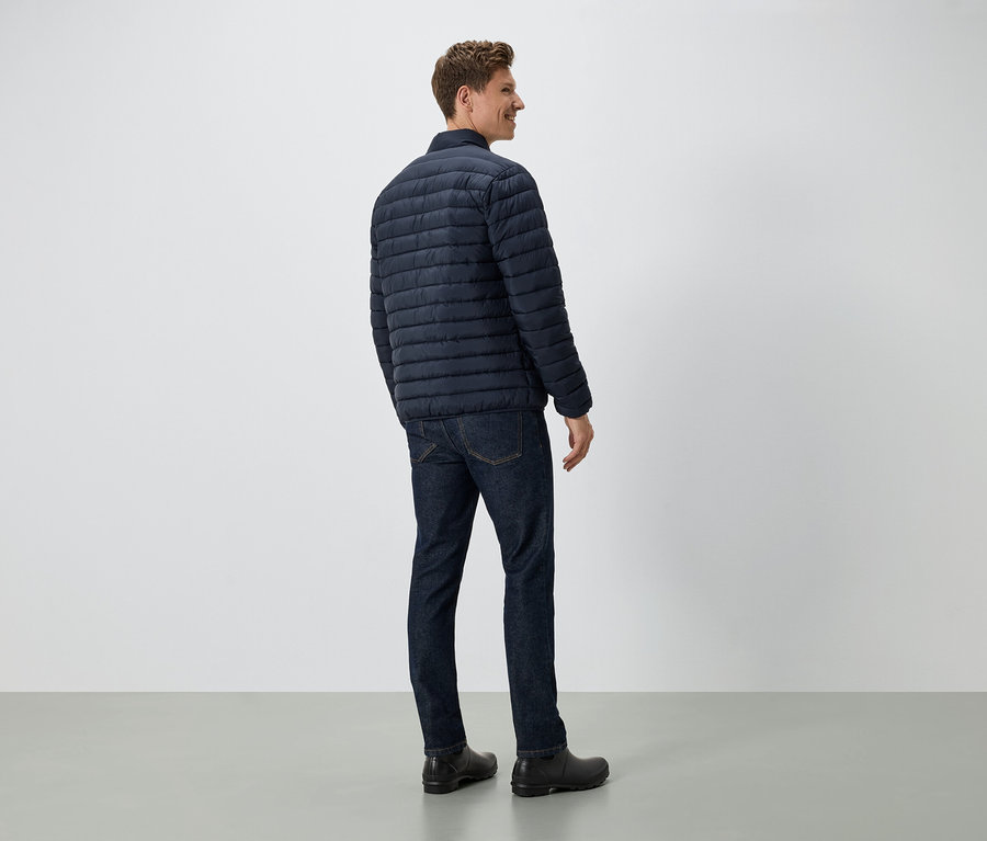 Muž stojí zády k fotoaparátu a má na sobě tmavě modrou péřovou bundu, džíny a černé boty.