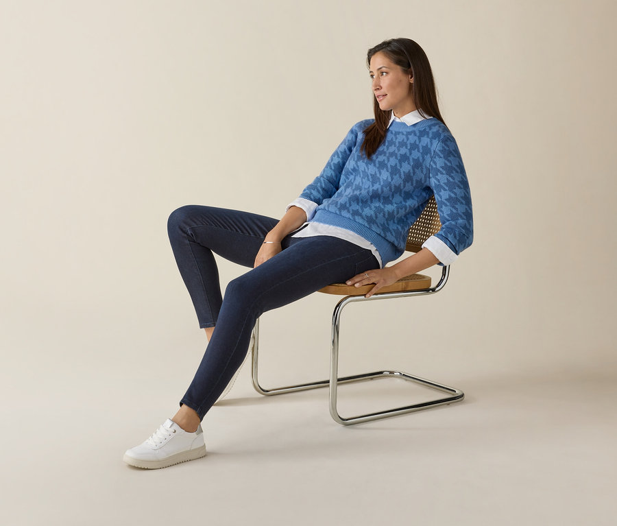 Žena sedí na židli a má na sobě modrý svetr a bílé tenisky Tom Tailor.