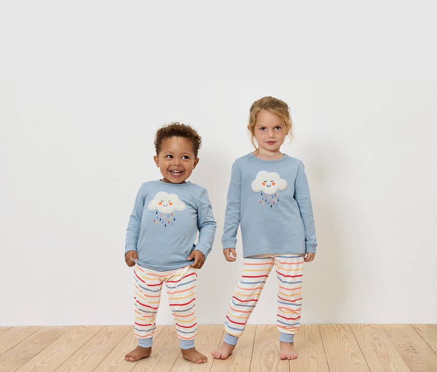 Dvě děti stojí na dřevěné podlaze a mají na sobě modrá trička s dlouhým rukávem a pruhované kalhoty.