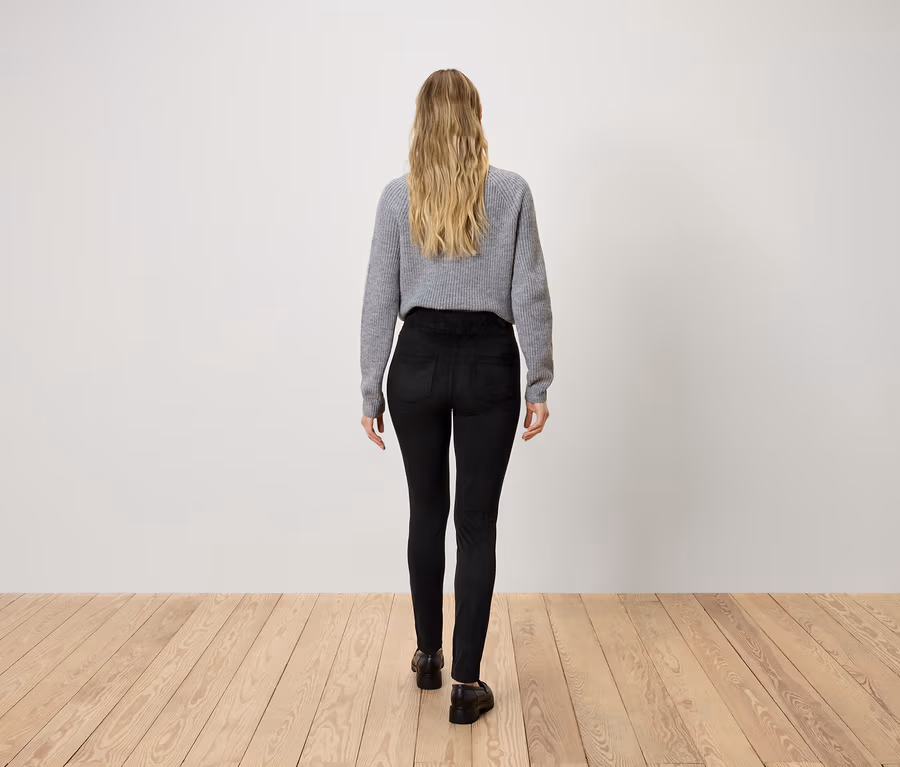 Žena s blond vlasy shora, oblečená do šedého svetru a černých kalhot, kráčí po dřevěné podlaze.