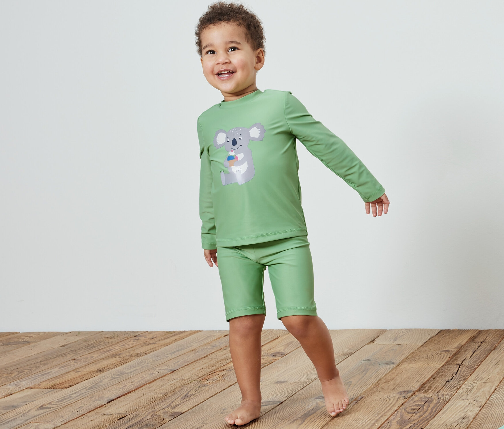 Chlapec v dětské soupravě s UV faktorem 80, koala, stojí na dřevěné podlaze vedle hračky. 