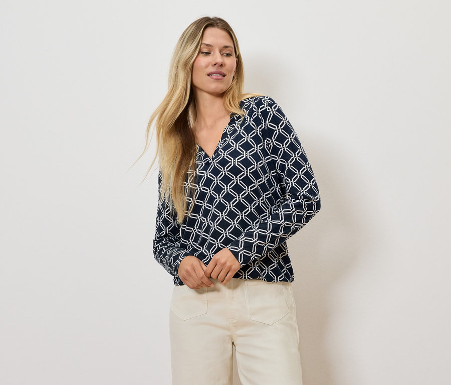 Blondýna pózuje v halence s geometrickým vzorem a světlých kalhotách.