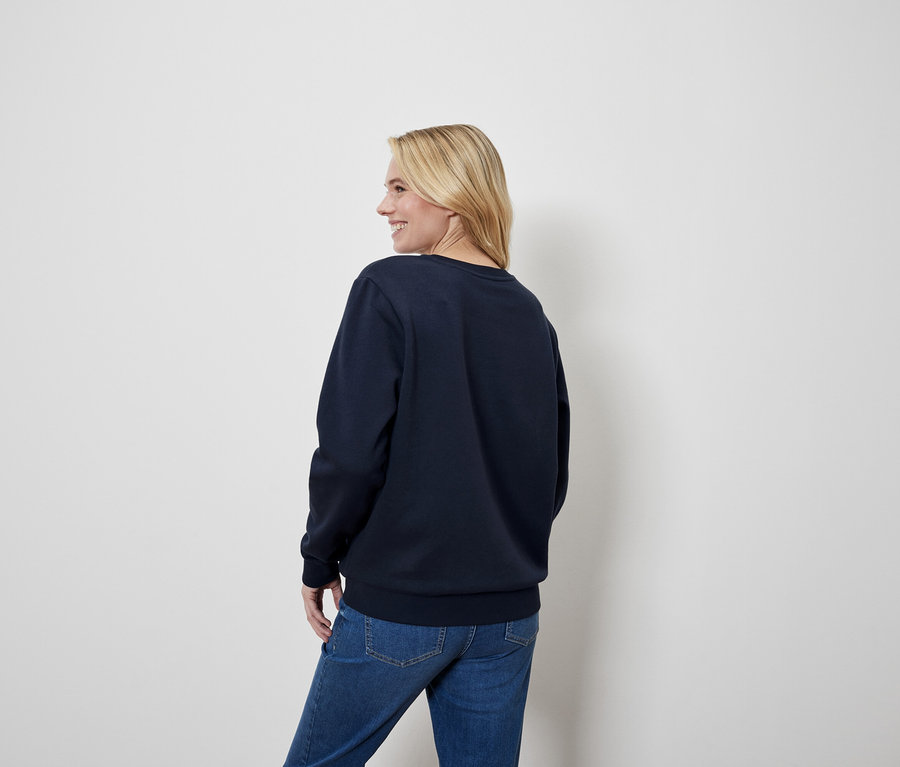 Blondýna se dívá přes rameno, má na sobě modrou mikinu a džíny.