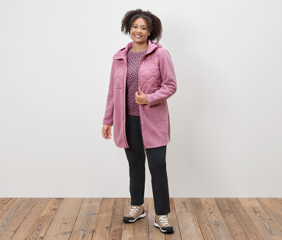 Žena v růžovém kabátě, vzorovaném topu a černých kalhotách stojí na dřevěné podlaze.