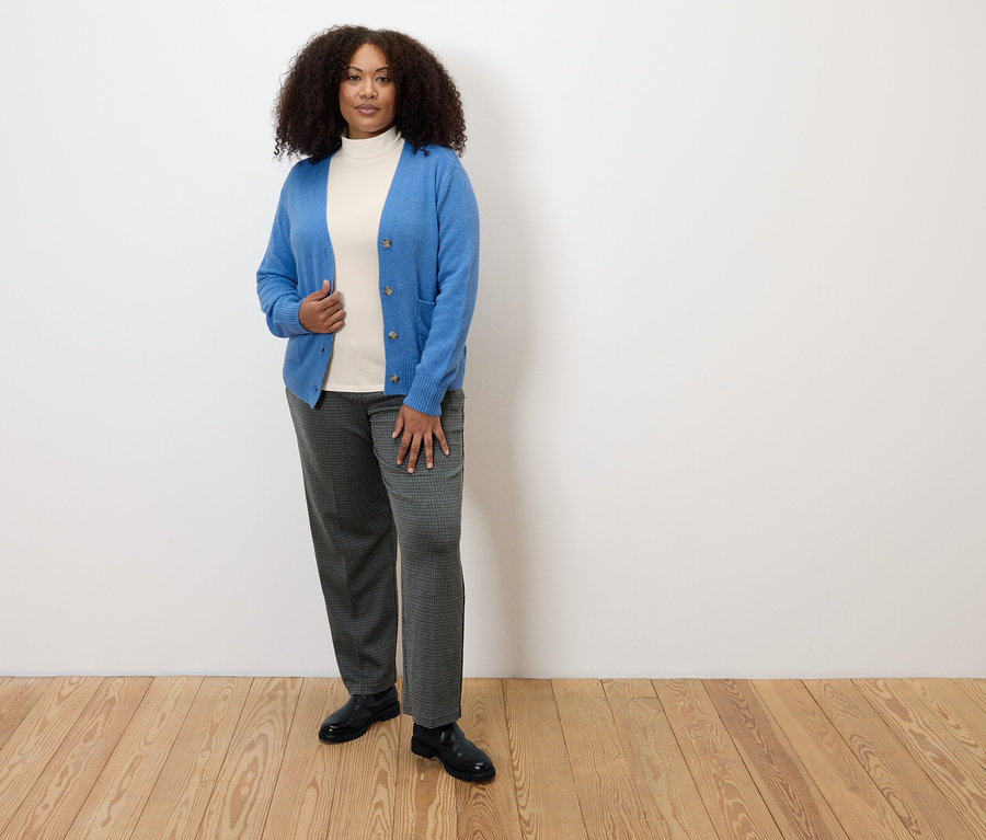 Žena stojící na dřevěné podlaze má na sobě kostkované pohodlné žerzejové kalhoty, bílý rolák a modrý kardigan.