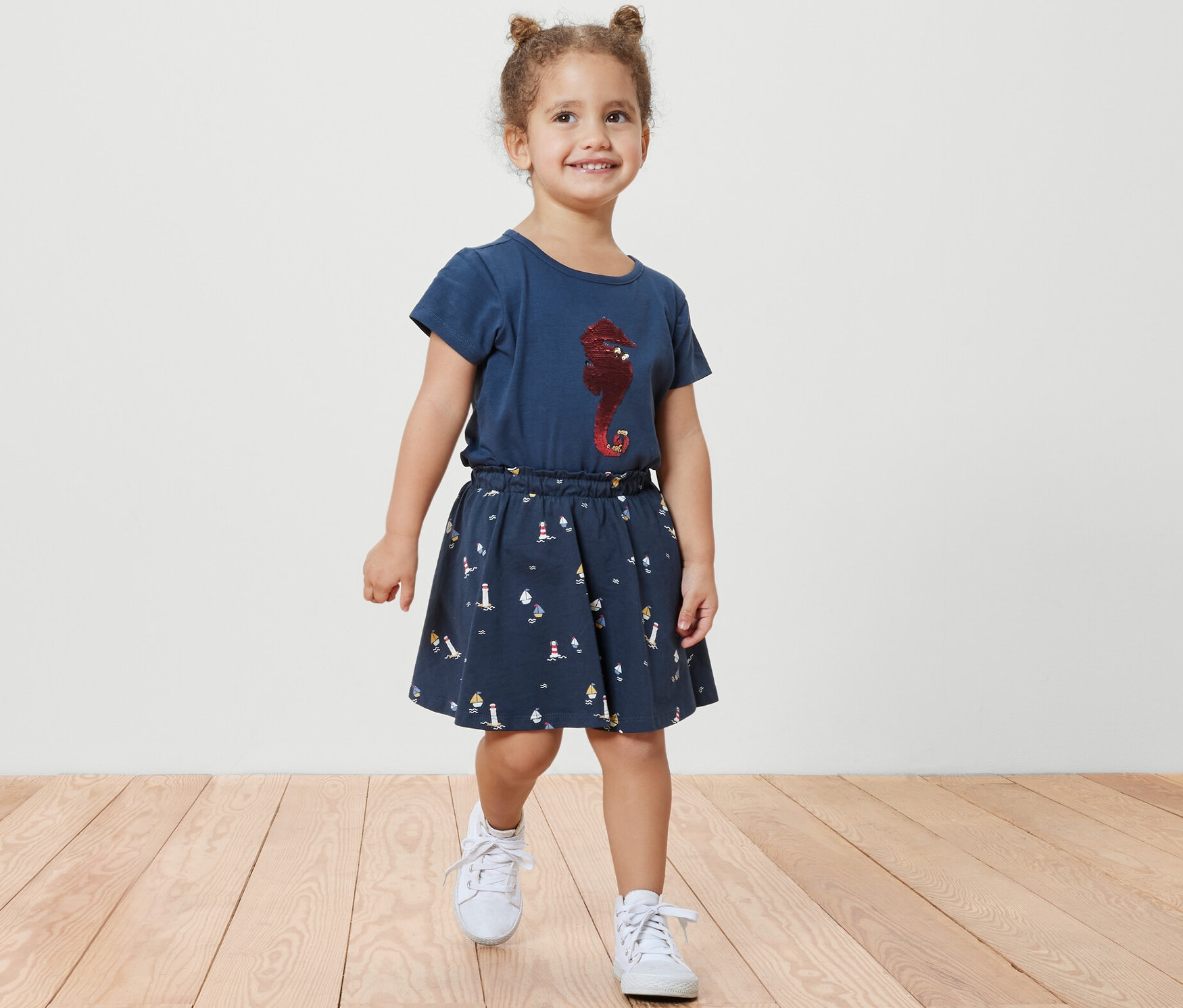 Usmívající se holčička kráčí po dřevěné podlaze a má na sobě dívčí žerzejovou sukni s šortkami s námořnickým vzorem a modré tričko s potiskem mořského koníka.
