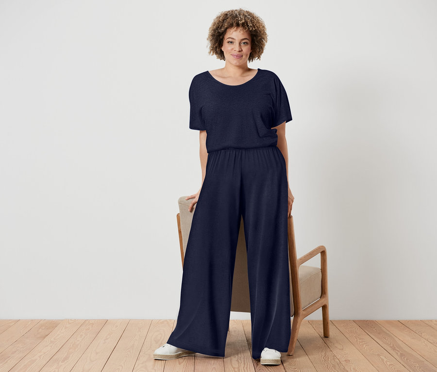 Žena s kudrnatými vlasy má na sobě tmavě modrý overal a bílé boty, stojí vedle židle s dřevěným rámem.