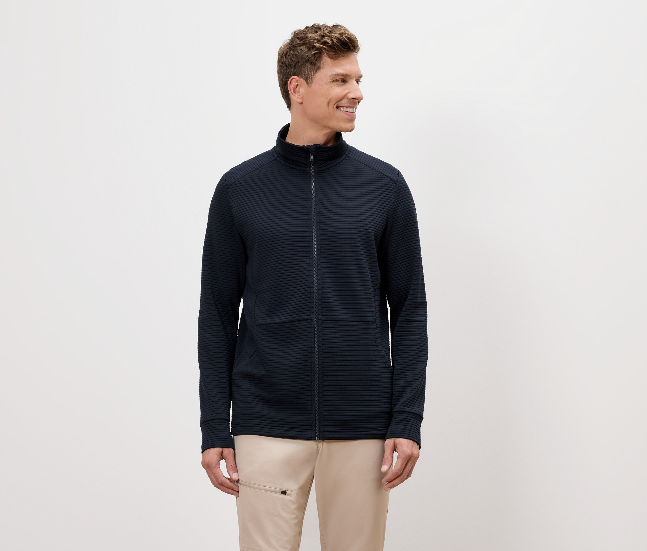 Muž s blond vlasy má na sobě tmavě modrou bundu na zip a béžové kalhoty.