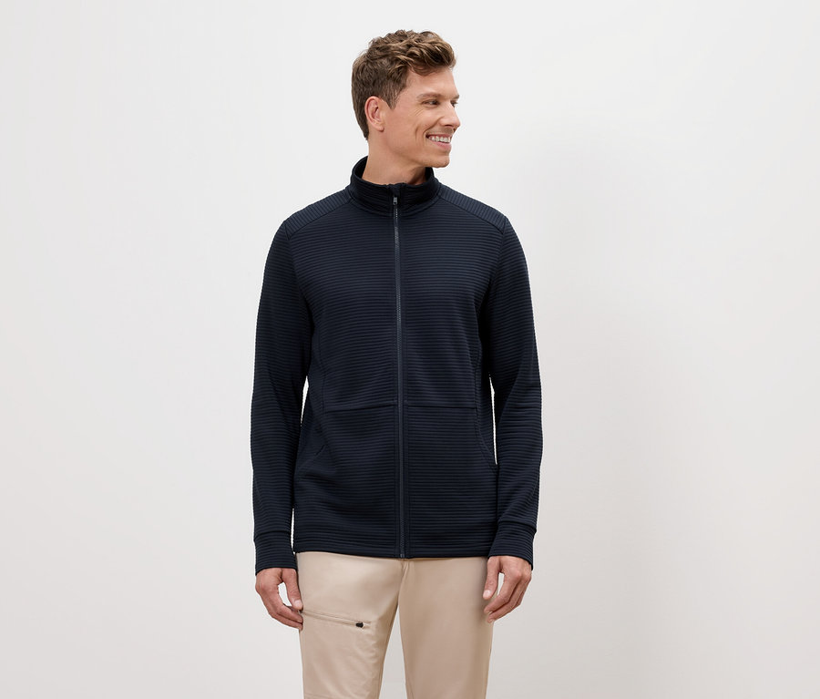 Muž s blond vlasy má na sobě tmavě modrou bundu na zip a béžové kalhoty.