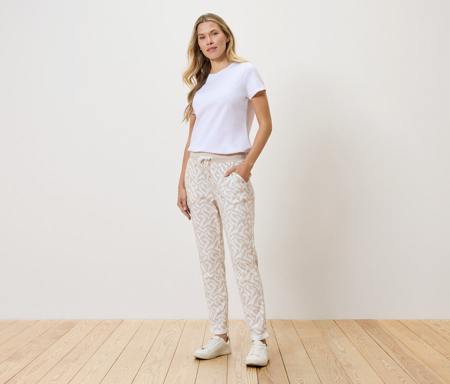 Žena stojí před stěnou, má na sobě bílé tričko, vzorované kalhoty a bílé tenisky.