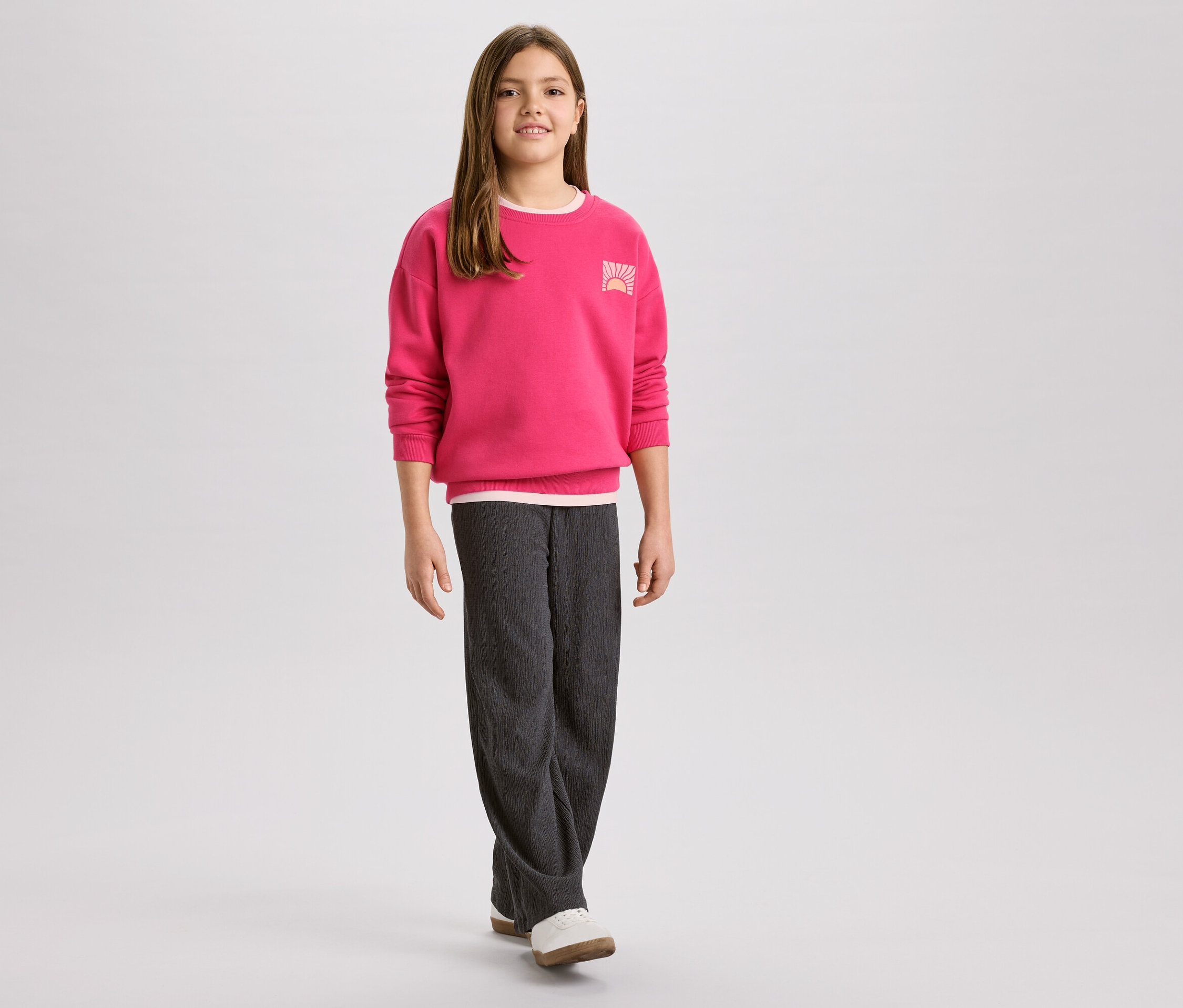 Dívka v růžové mikině s logem východu slunce a šedých manšestrových kalhotách, k tomu bílé boty.